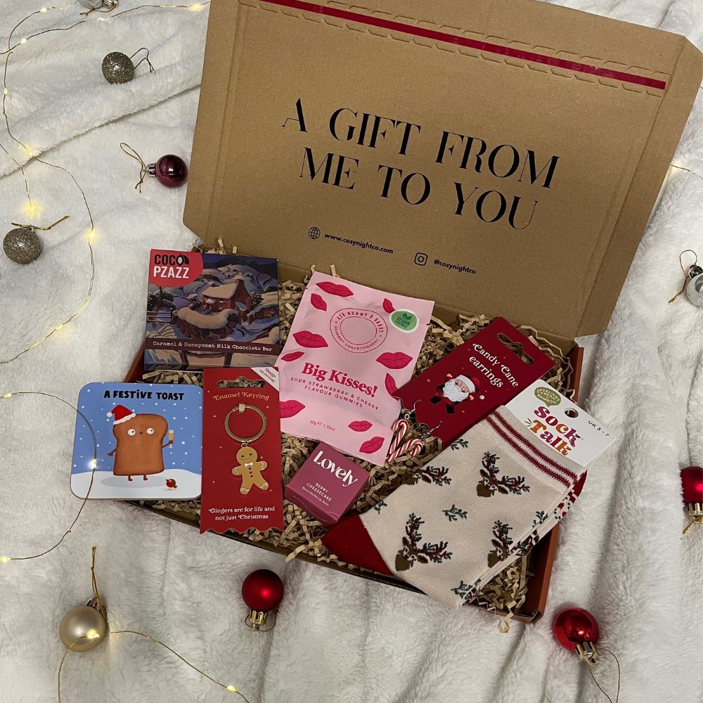 A Christmas themed gift box filled with cosy treats including chocolate, festive socks, earrings, a coaster, and a face mask, displayed on a white blanket with Christmas lights and baubles.