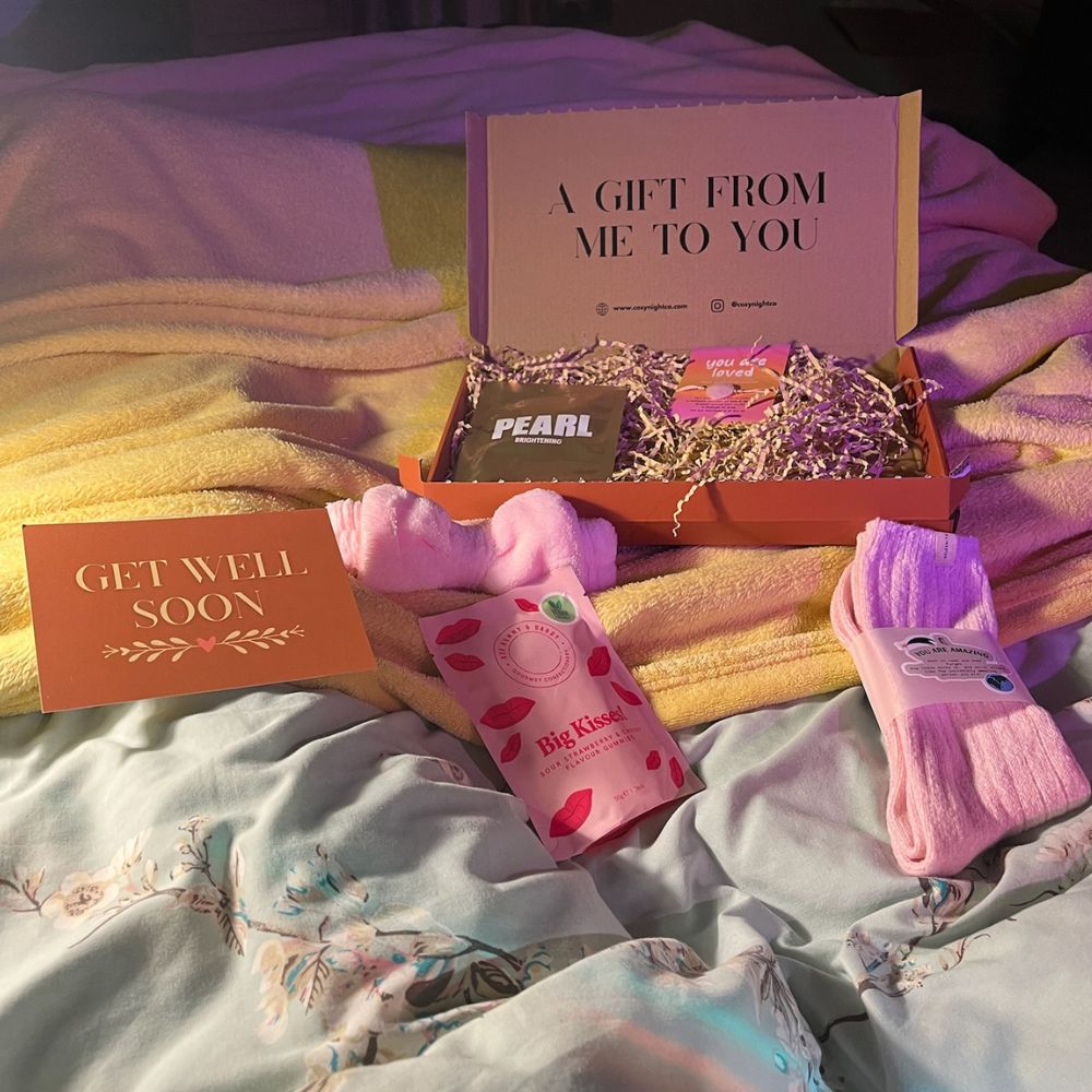 A “Get Well Soon” letterbox gift box on a bed with pink cosy socks, a pearl face mask, sweets, a headband, and a cheerful message card.
