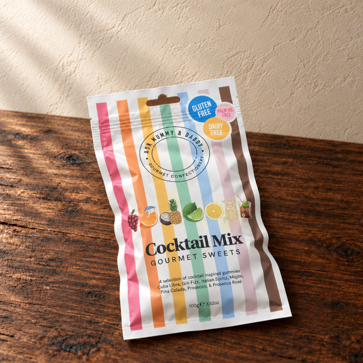 A share bag of 'Cocktail Mix Gourmet Sweets', Featuring colourful vertical stripes and illustrations of various fruits, resting on a dark wooden surface against a textured beige wall.