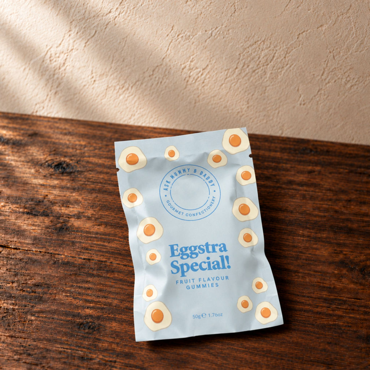 A pouch of Eggstra Special fruit flavour gummies placed on a dark wooden tabletop against a textured beige wall. The pouch is light blue with illustrations of fried eggs and blue text.