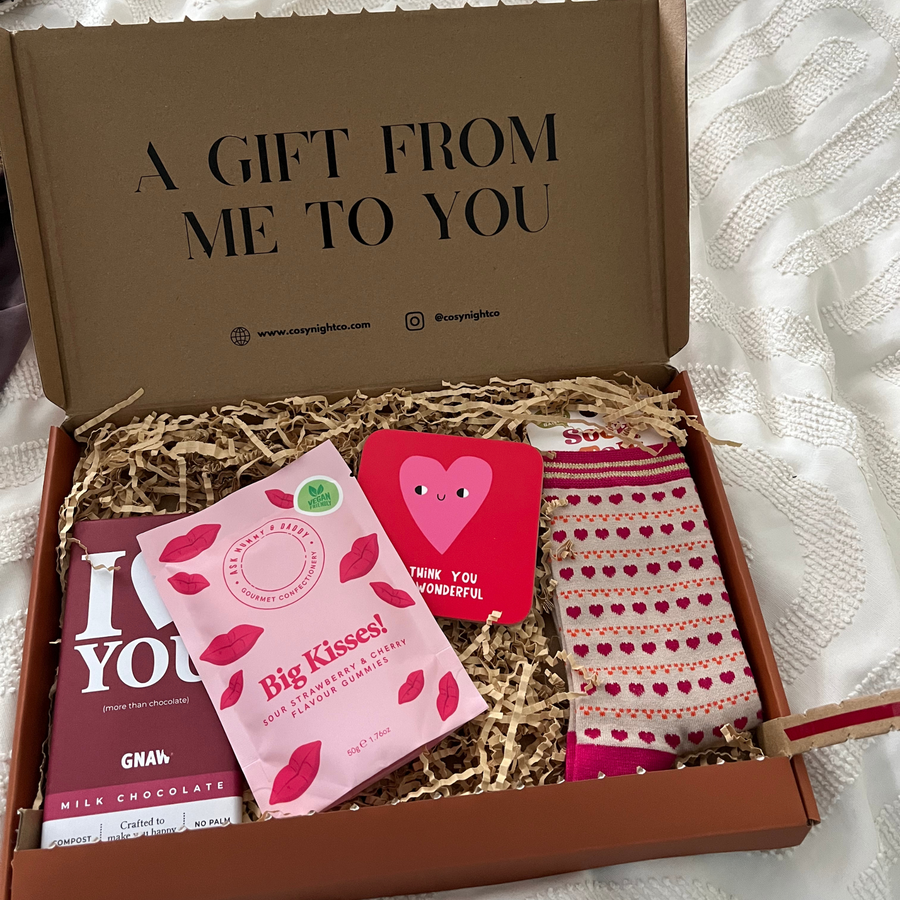 Gift box with 'A Gift From Me To You' text, including chocolate, sweets, coaster, and socks on kraft shredded paper.