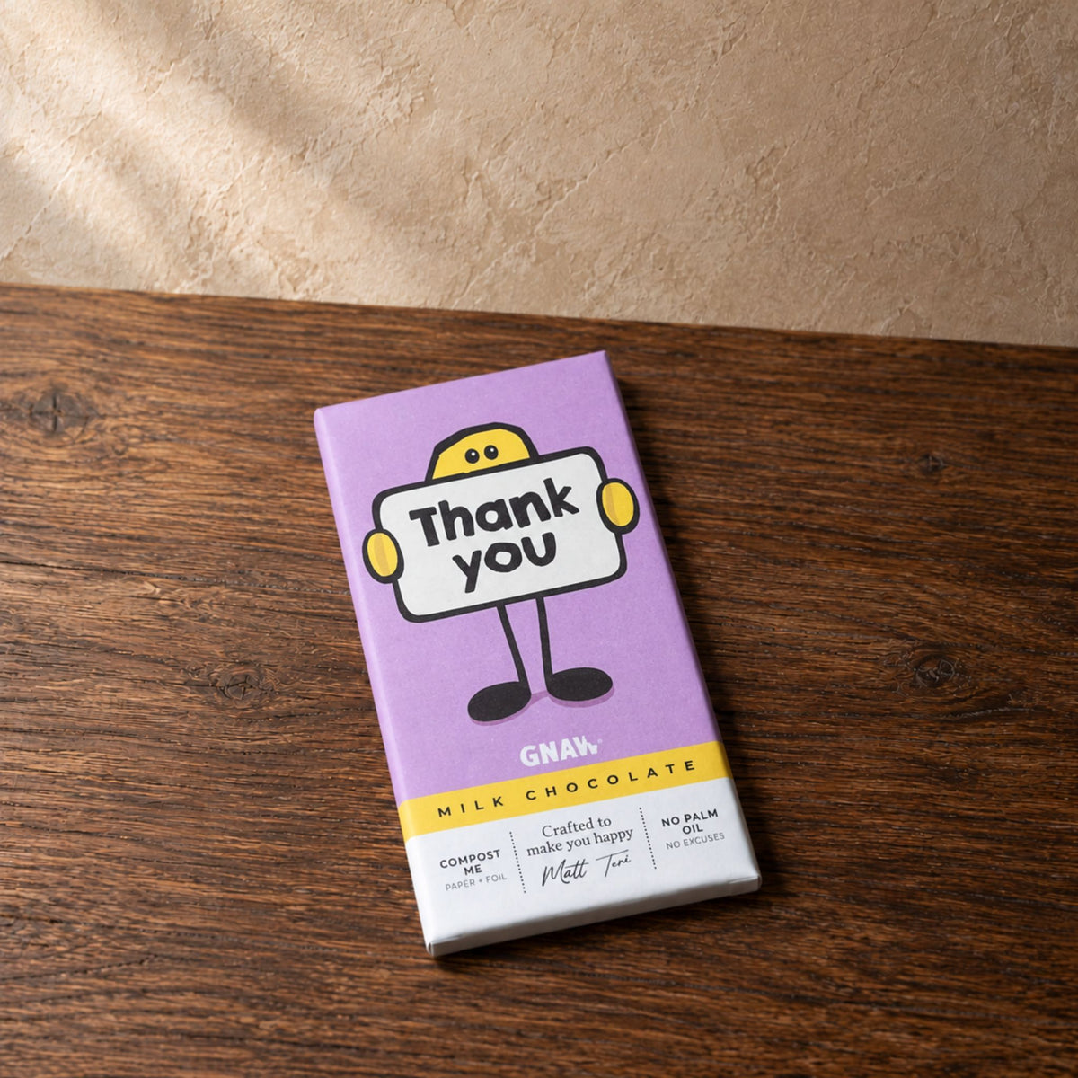 A purple milk chocolate bar wrapper labeled 'Thank you' with a cartoon character, placed on a dark wooden tabletop against a textured beige wall.