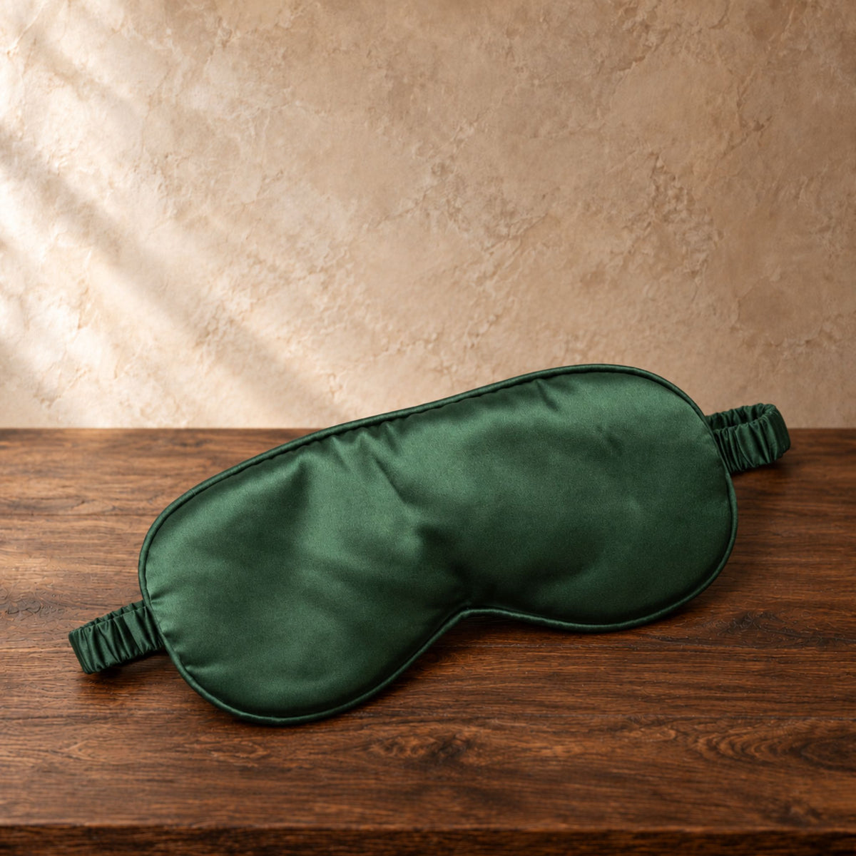 A dark green satin sleep mask with elastic straps, resting on a dark wooden surface against a textured beige wall.