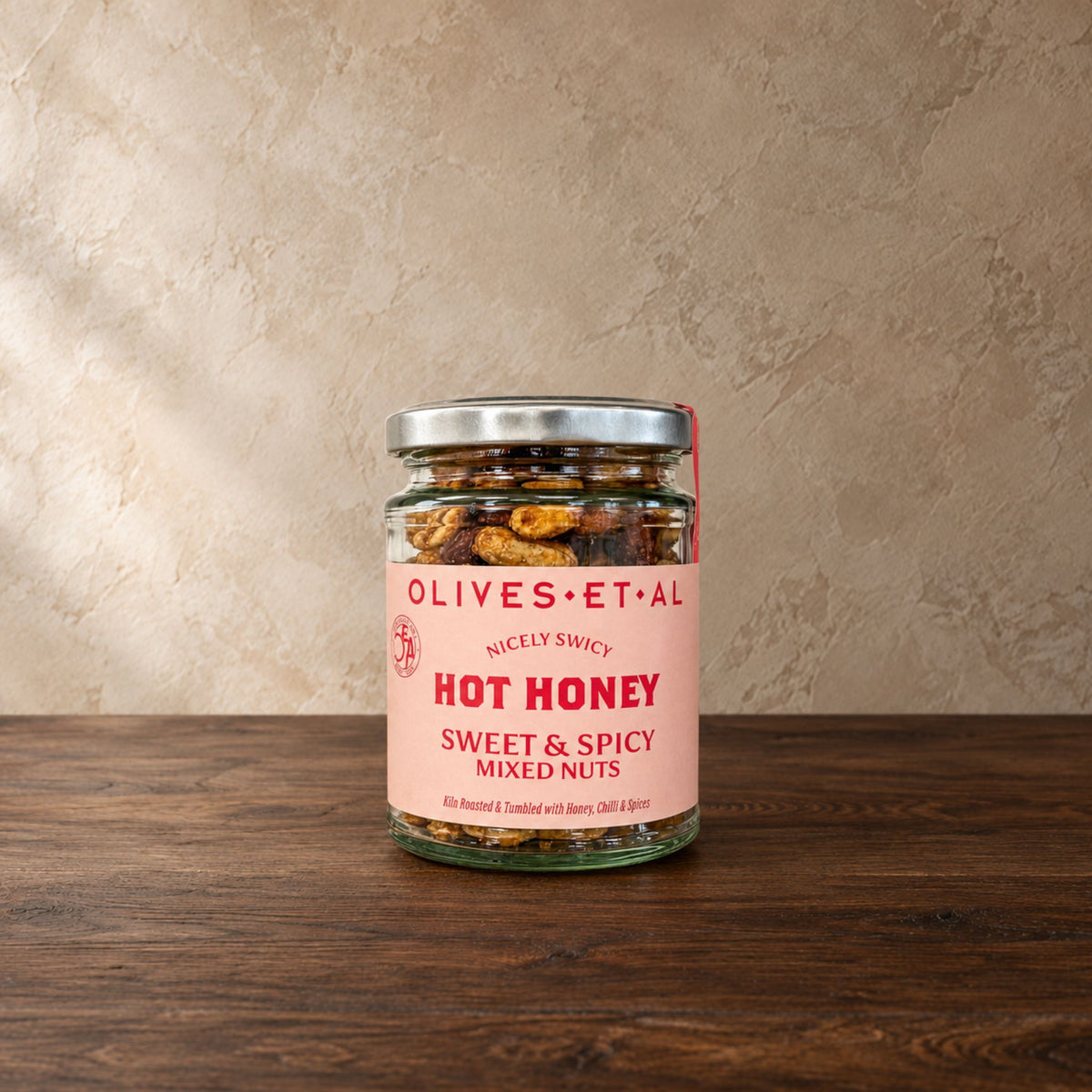 A glass jar of Olives Et Al 'Hot Honey' Sweet & Spicy Mixed Nuts sits on a dark wooden table against a textured beige wall. The jar has a silver metal lid and a light pink label with red text.