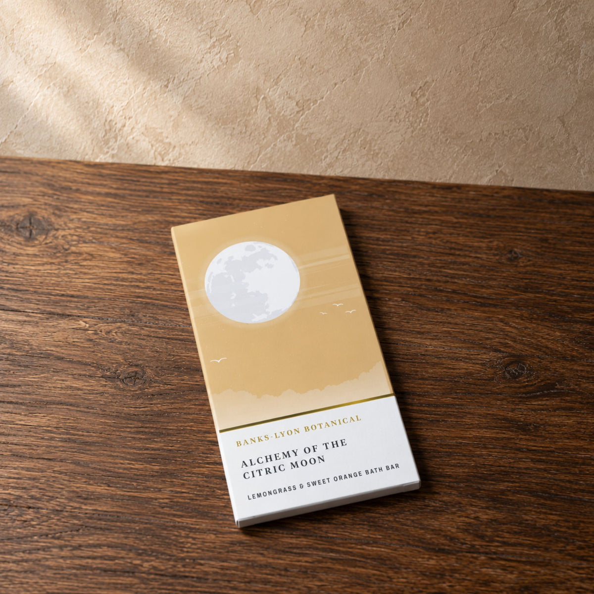 A rectangular bath bar package labeled 'Alchemy of the Citric Moon' rests on a dark wood surface against a textured beige wall, with soft natural light casting shadows across the scene.