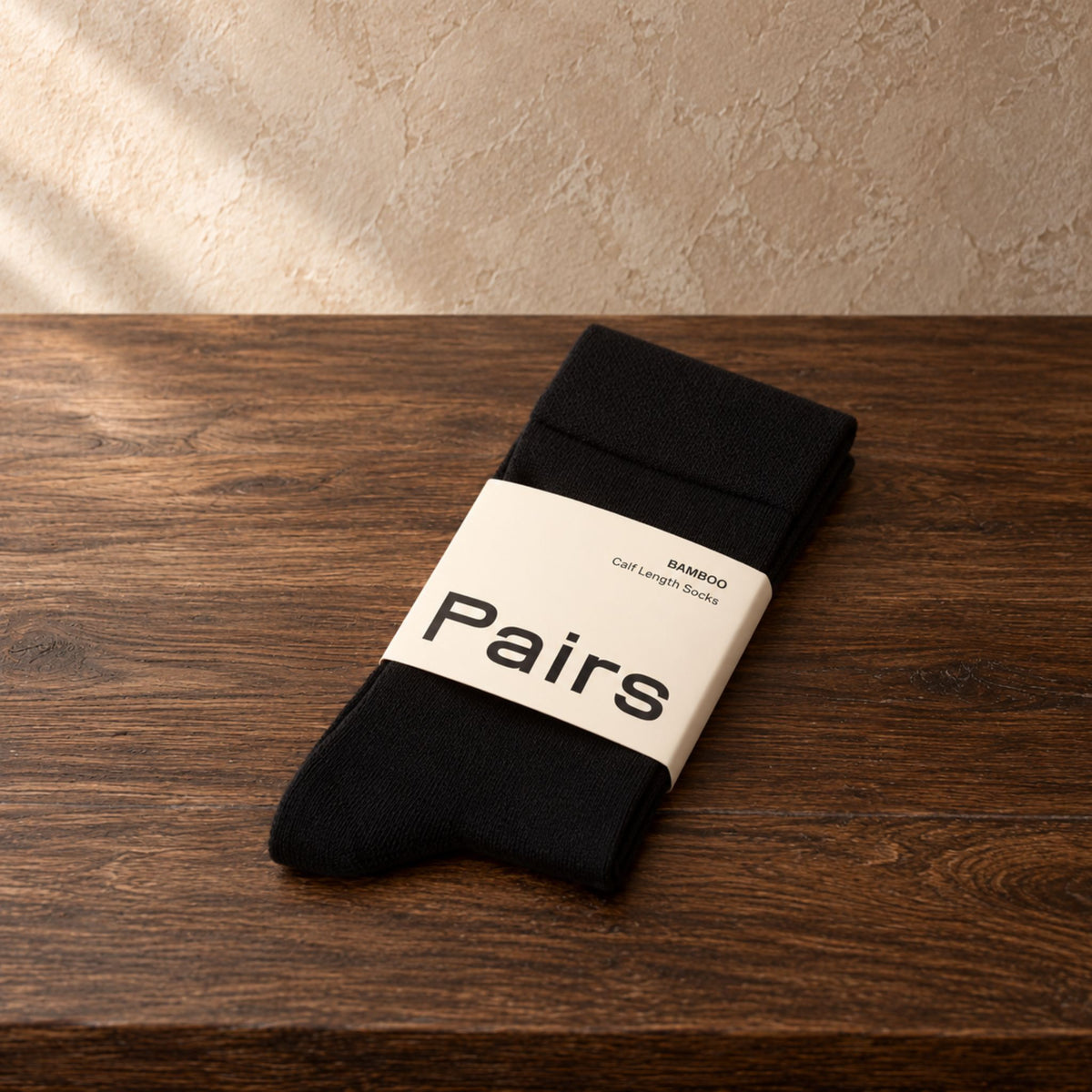 A pair of black socks wrapped in a white paper band labeled 'Pairs' and 'BAMBOO Calf Length Socks', resting on a dark wooden surface against a textured beige wall.