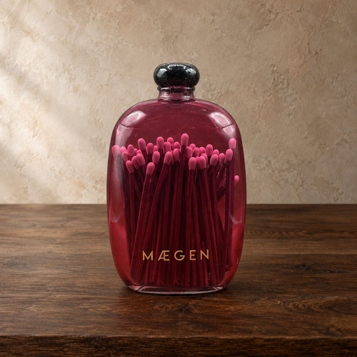 A pink glass bottle with a black cap, filled with pink matchsticks, sitting on a dark wooden table against a textured beige wall. The brand name is printed in gold on the front of the bottle.