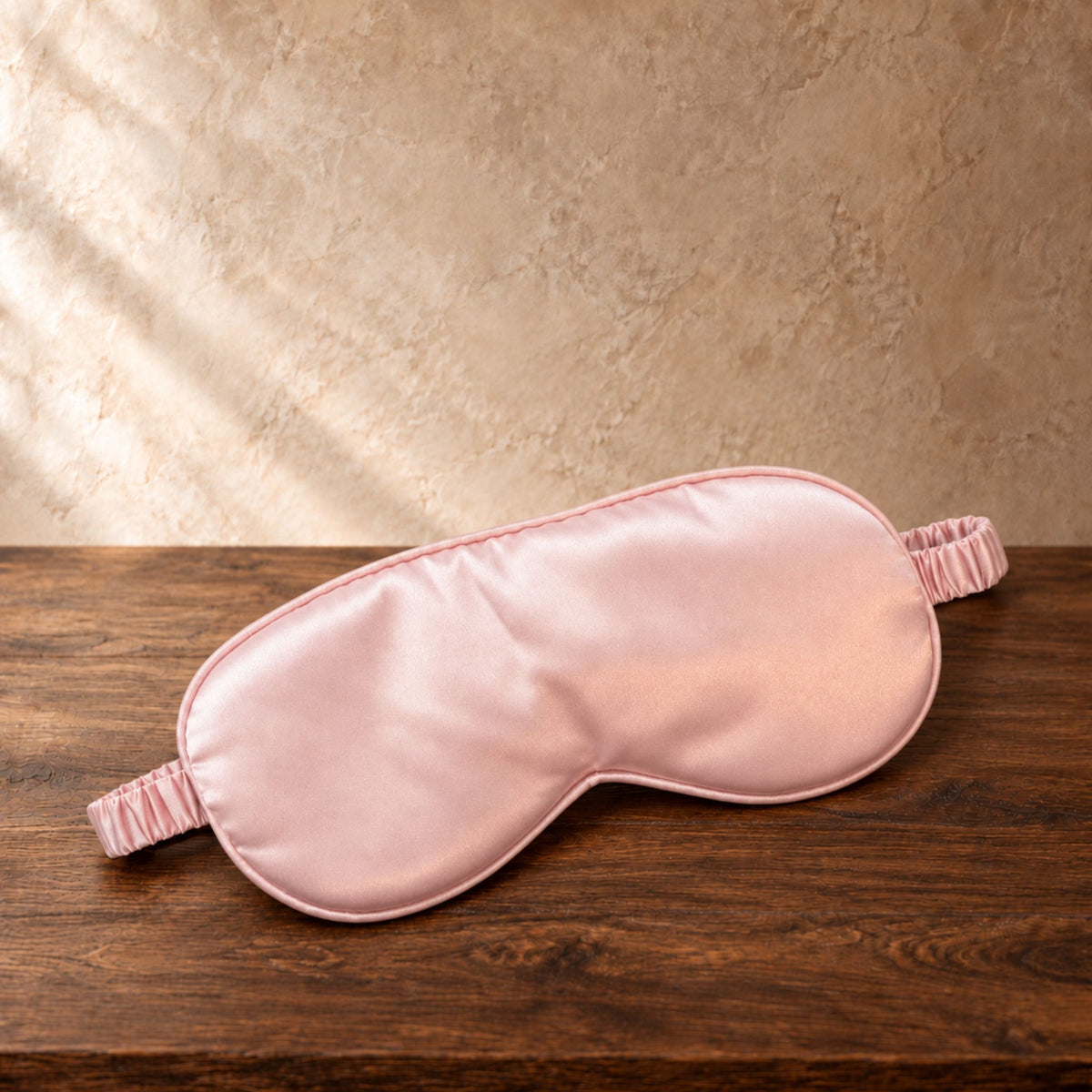 A light pink satin sleep mask with matching elastic ear loops, resting on a dark brown wooden surface against a textured beige wall with soft diagonal light rays.