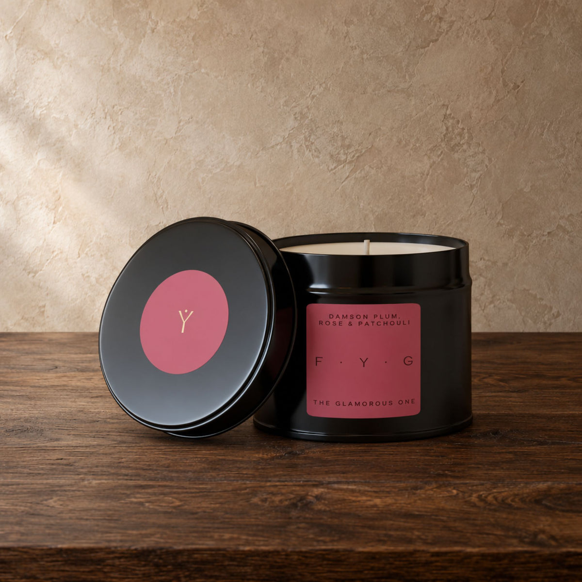 A black metal candle tin with its lid leaning beside it on a dark wooden surface. The candle has a pink label reading 'Garden Plum Rose & Patchouli'.