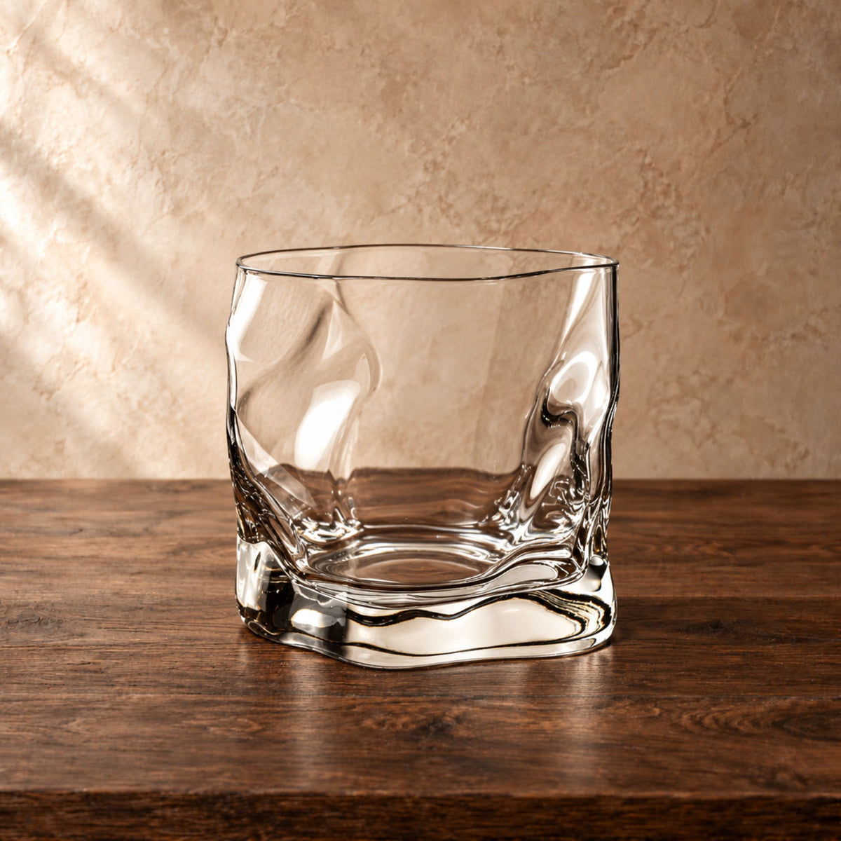A clear glass whiskey tumbler with a wavy, textured organic pattern, sitting on a dark wooden table against a textured beige wall.