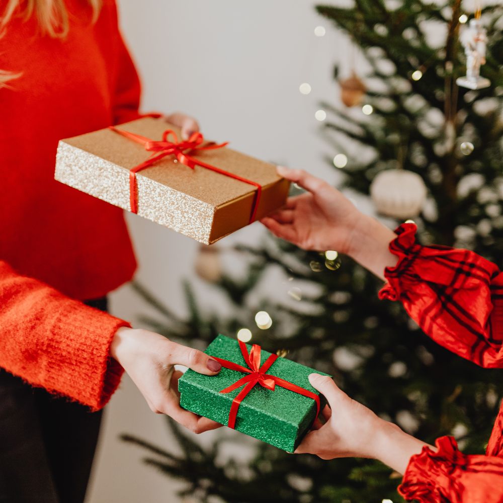 Two people exchanging wrapped Christmas gifts beside a decorated tree, with red and green present boxes tied with ribbon.