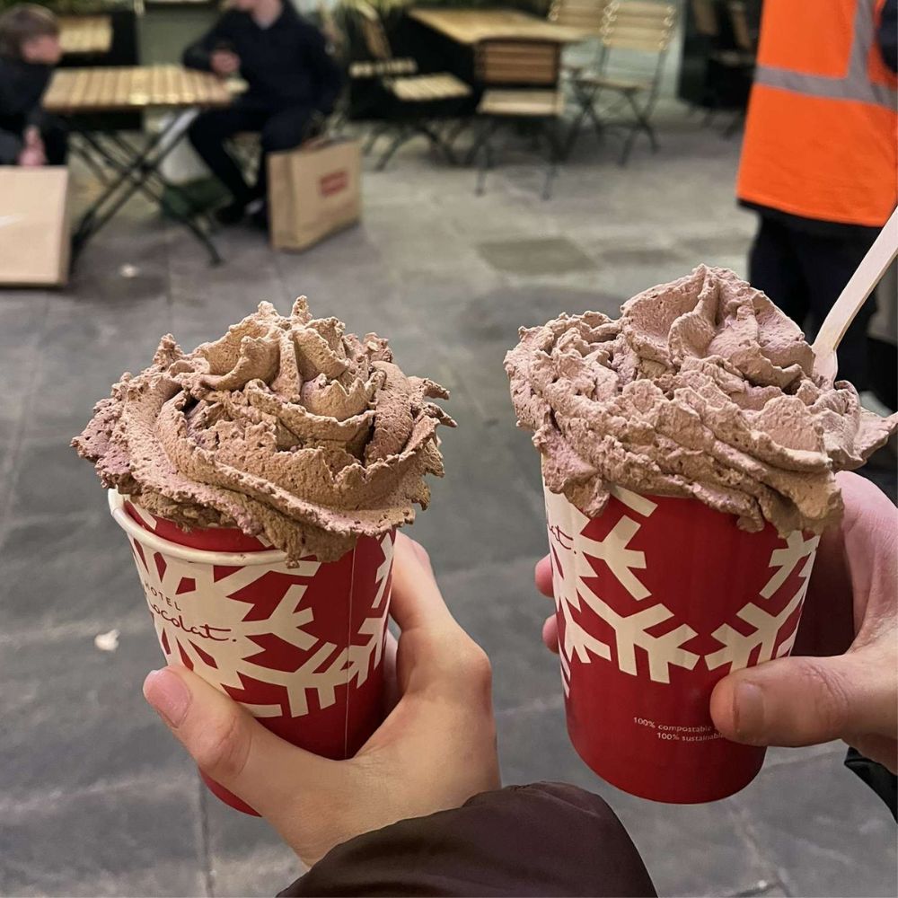 Two hands holding Christmas-themed red hot chocolate cups with chocolate whipped cream on top outside, with outdoor seating and shopping bags in the background.