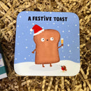 Cute Christmas coaster featuring a smiling slice of toast wearing a Santa hat and holding a drink in the snow. With the writing 'A Festive Toast' at the top.