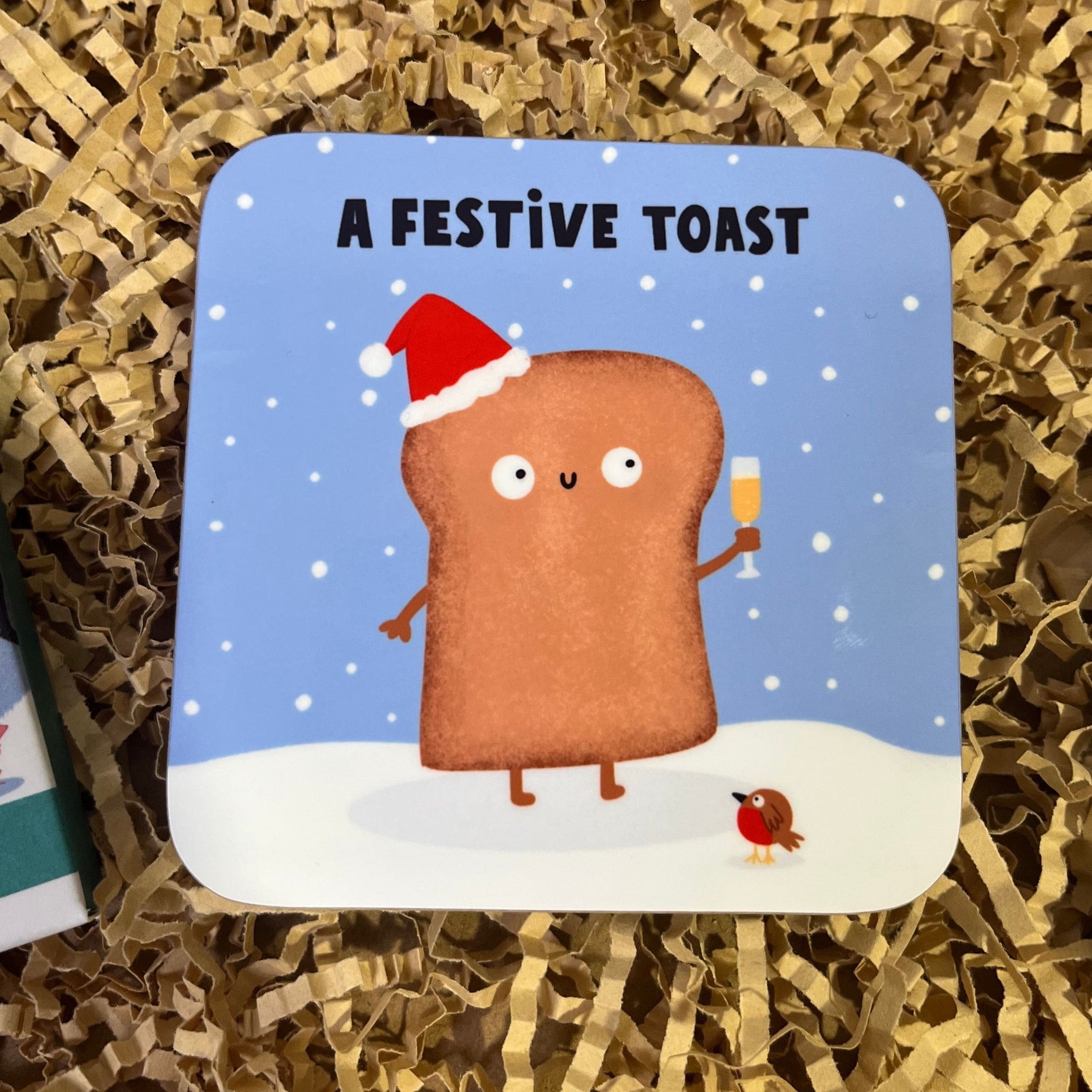 Cute Christmas coaster featuring a smiling slice of toast wearing a Santa hat and holding a drink in the snow. With the writing 'A Festive Toast' at the top.