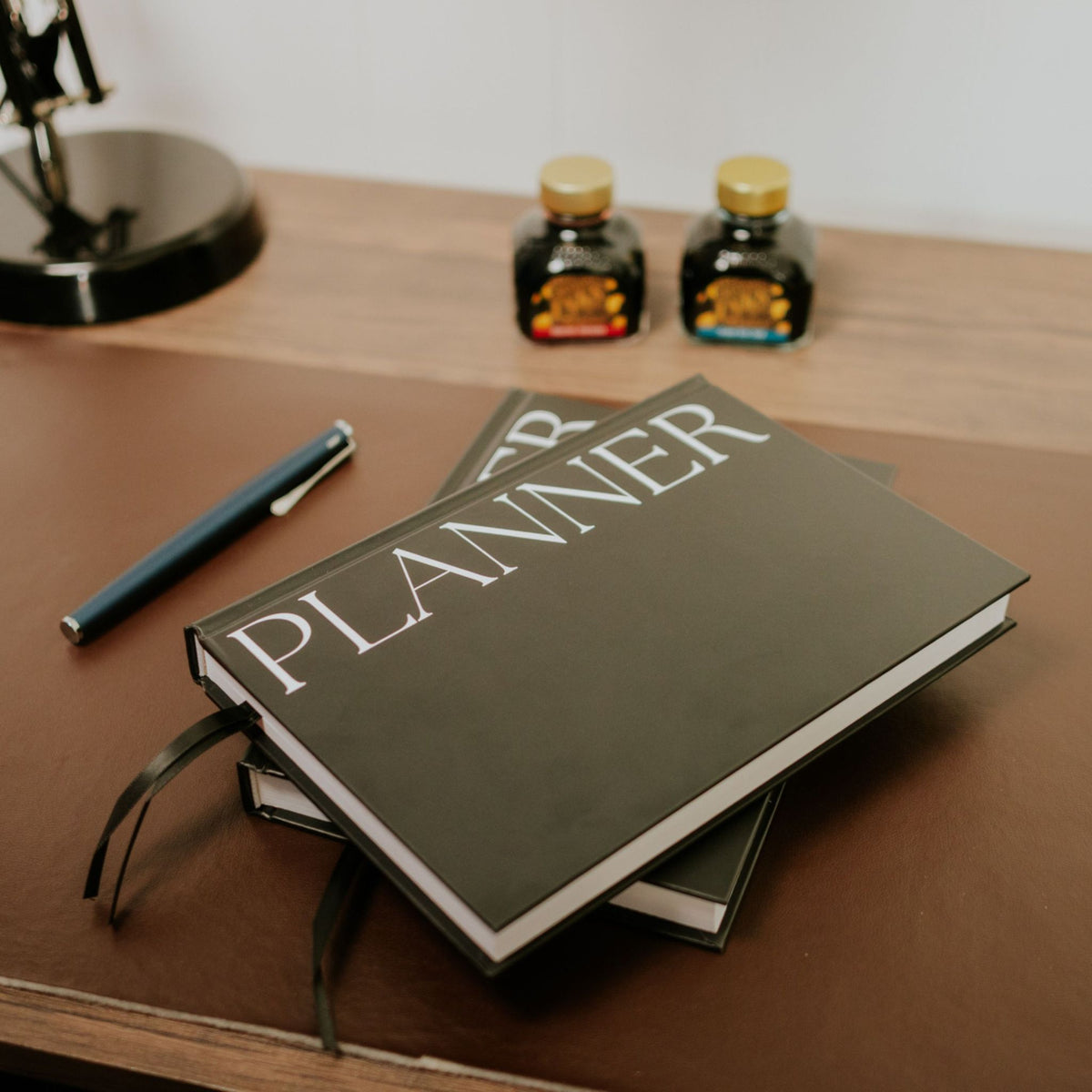 A black hardcover planner with large white 'PLANNER' text on the cover, stacked on another planner on a wooden desk, with a blue pen nearby and two small ink bottles in the background.