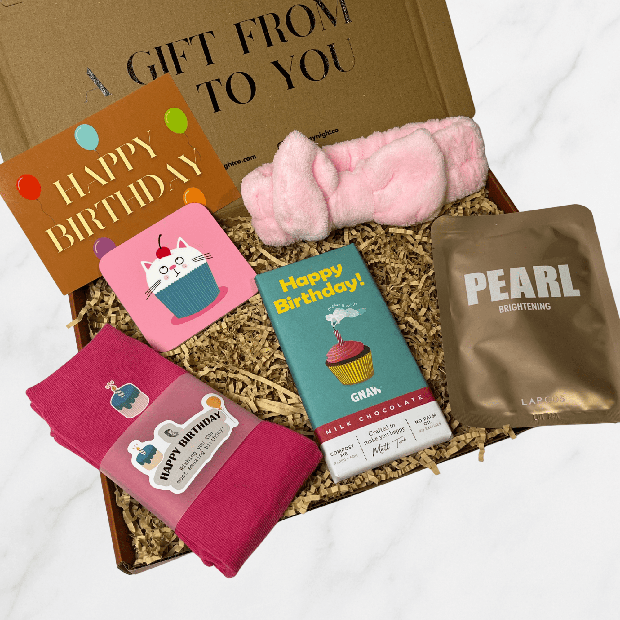 Birthday gift box with pink happy birthday themed socks, cat cupcake coaster, Happy Birthday chocolate bar, pearl sheet mask, and pink spa headband.