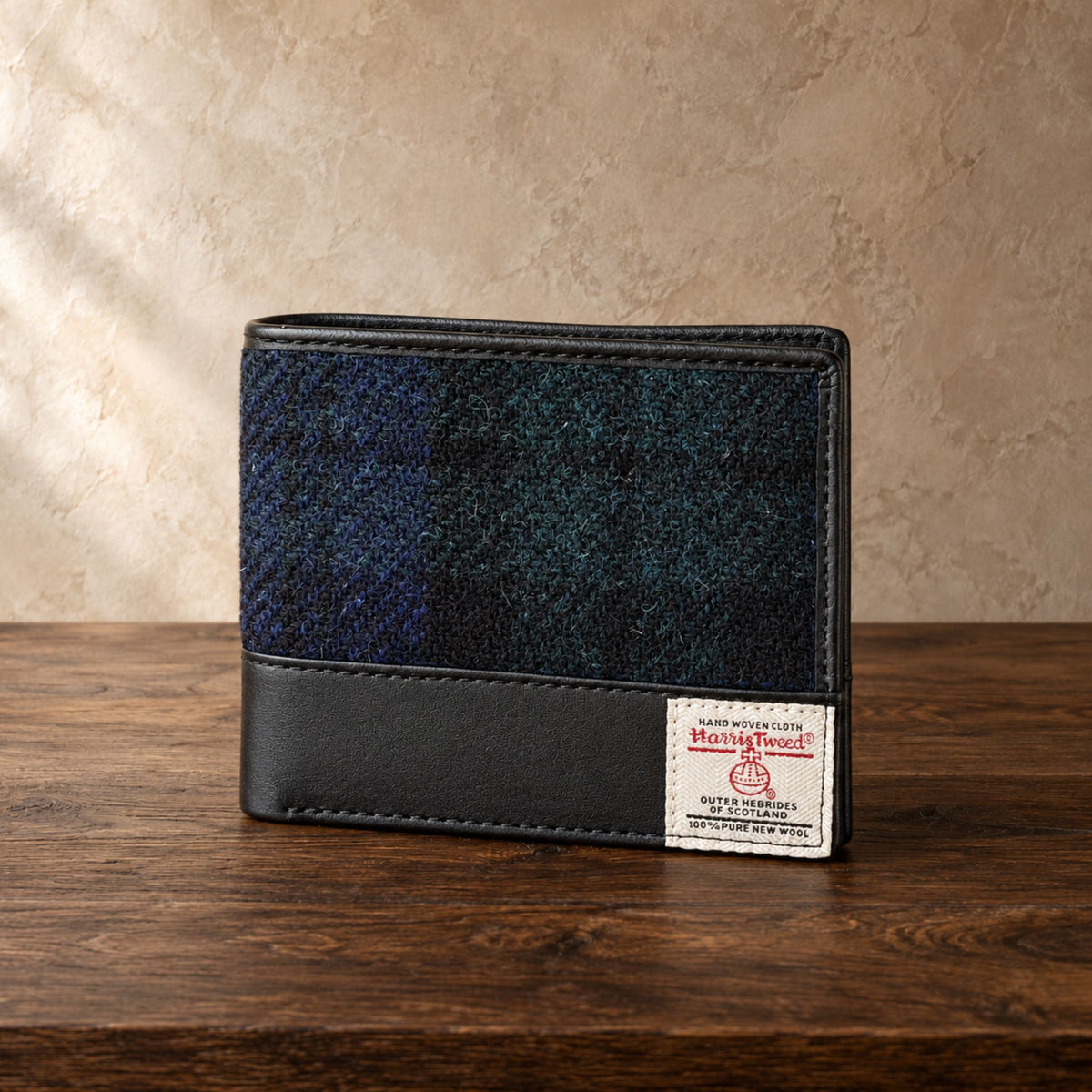 A wallet made of dark blue and black plaid tweed with a black leather lower section, resting on a dark wooden table against a textured beige wall.