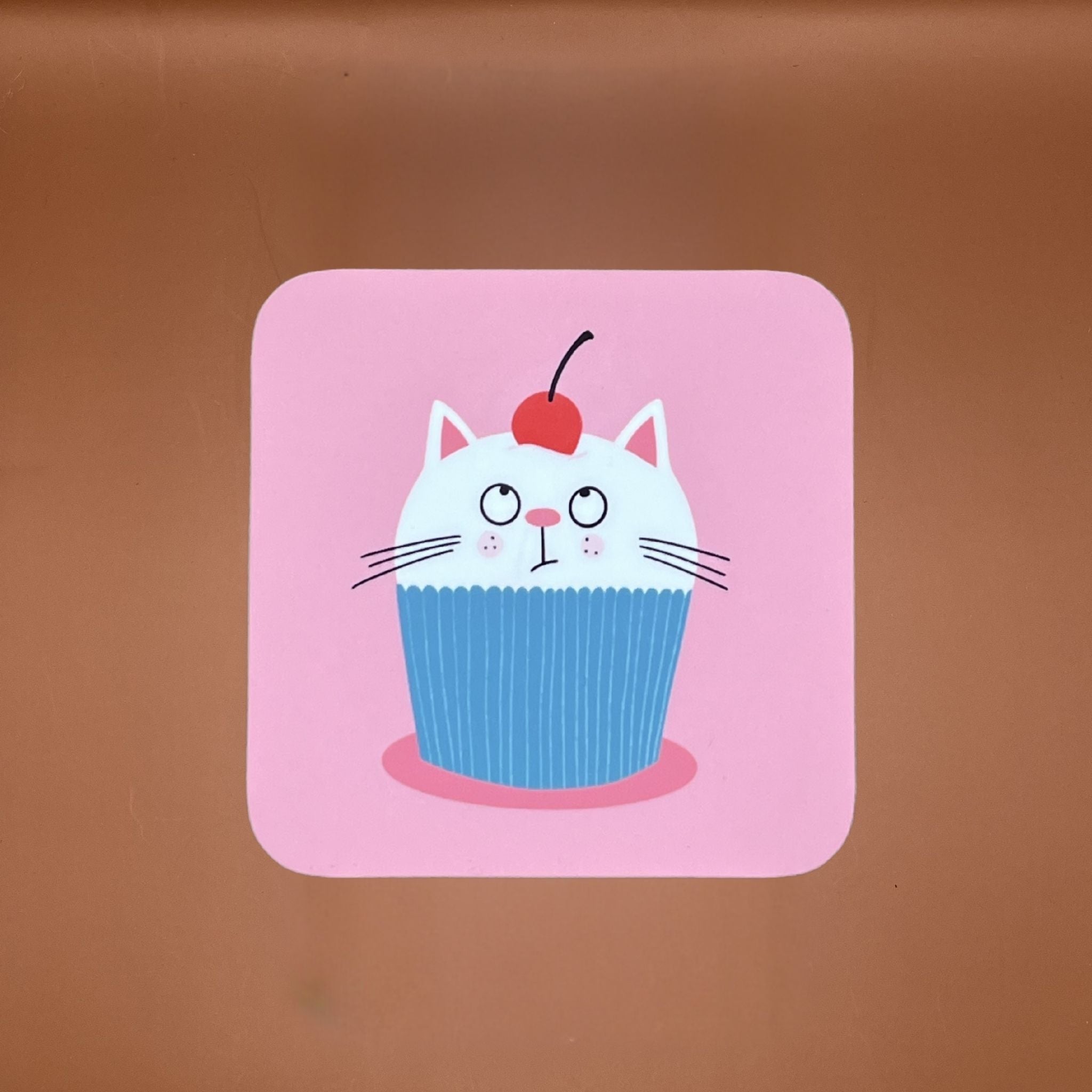 Pink coaster featuring a cute illustration of a white cat inside a green cupcake wrapper with a red cherry on its head.