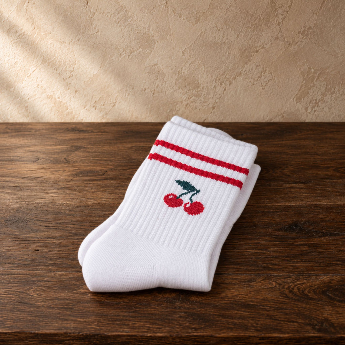 A pair of white ribbed crew socks folded on a dark wooden surface, featuring two red horizontal stripes near the cuff and a small embroidered red cherries..