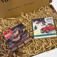 Two festive Coco Pzazz chocolate bars, one caramel and honeycomb and one milk chocolate with snow flurries, displayed in a Christmas gift box.