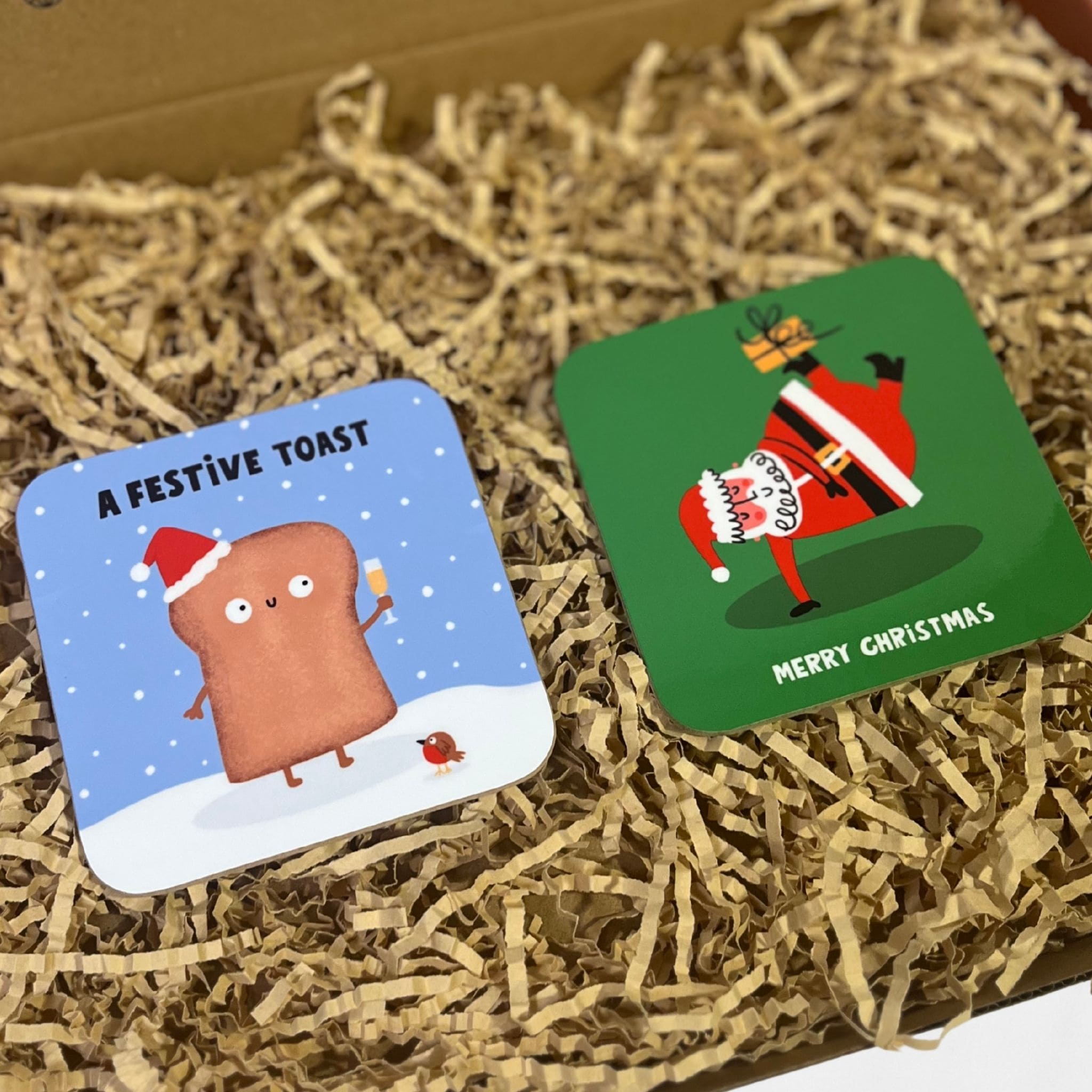 Two festive coasters, one featuring a cheerful toast character holding a drink in the snow, and another showing Santa balancing on one hand with a gift.