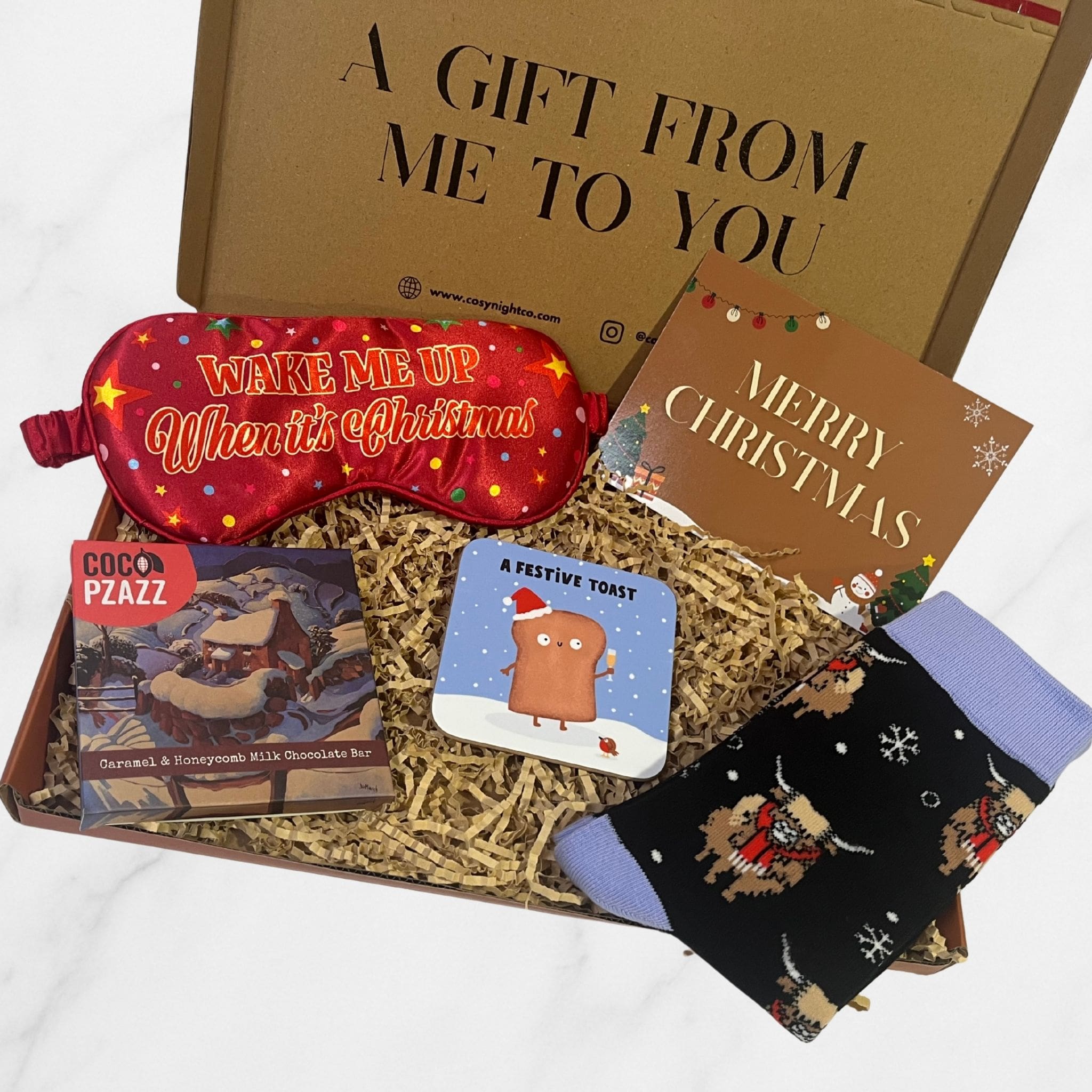 Festive letterbox Christmas gift box containing a red “Wake Me Up When It’s Christmas” sleep mask, Coco Pzazz chocolate bar, Highland cow socks, coaster, and Merry Christmas card.