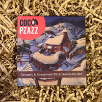 Coco Pzazz Caramel & Honeycomb Milk Chocolate Bar featuring a snowy cottage illustration on festive packaging nestled in shredded paper.