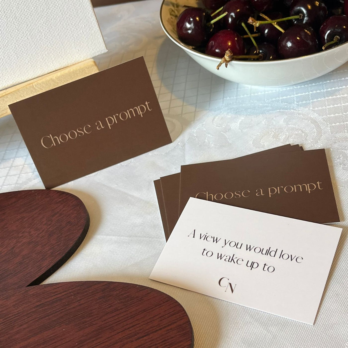 Prompt cards displayed on a white tablecloth, featuring one dark brown card reading 'Choose a prompt' and a white card with a custom quote. A white bowl of fresh cherries and wooden paint palette next to them.