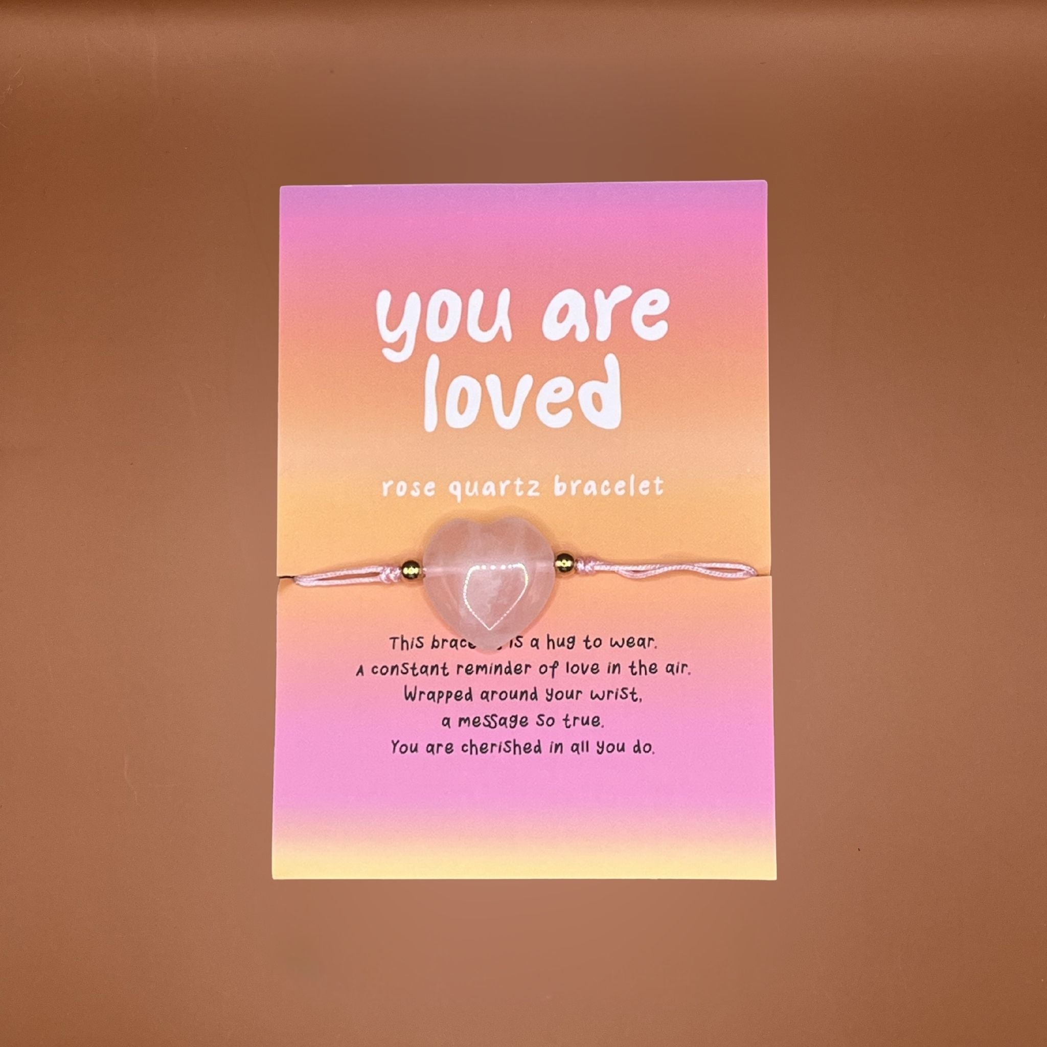 Rose quartz heart bracelet with 'you are loved' card on a sunset gradient background