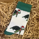 White Christmas socks with green cuffs featuring turtles wearing Santa hats and scarves, displayed on shredded paper packaging inside a gift box.