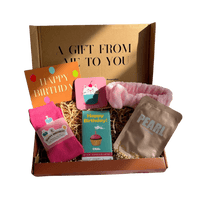 Gift box filled with a pair of socks, a facemask, a cat cupcake coaster, chocolate, a pink fluffy headband, and a message card saying Happy Birthday.