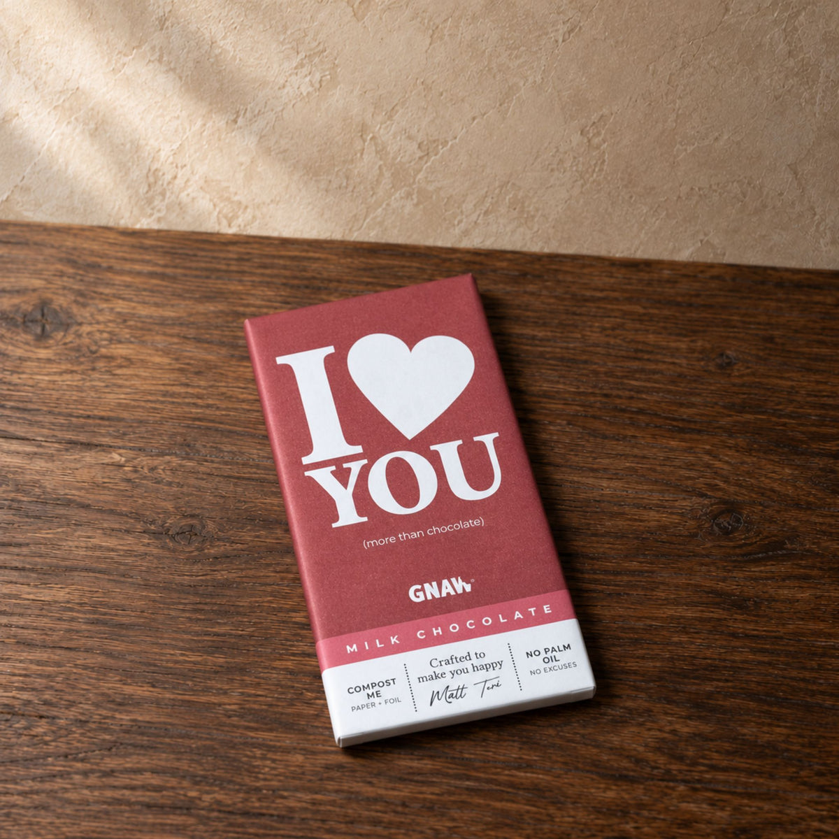 A milk chocolate bar package with a dusty rose red wrapper and white text reading 'I [heart] YOU' and 'GNAW', placed on a dark wood tabletop against a textured beige wall.