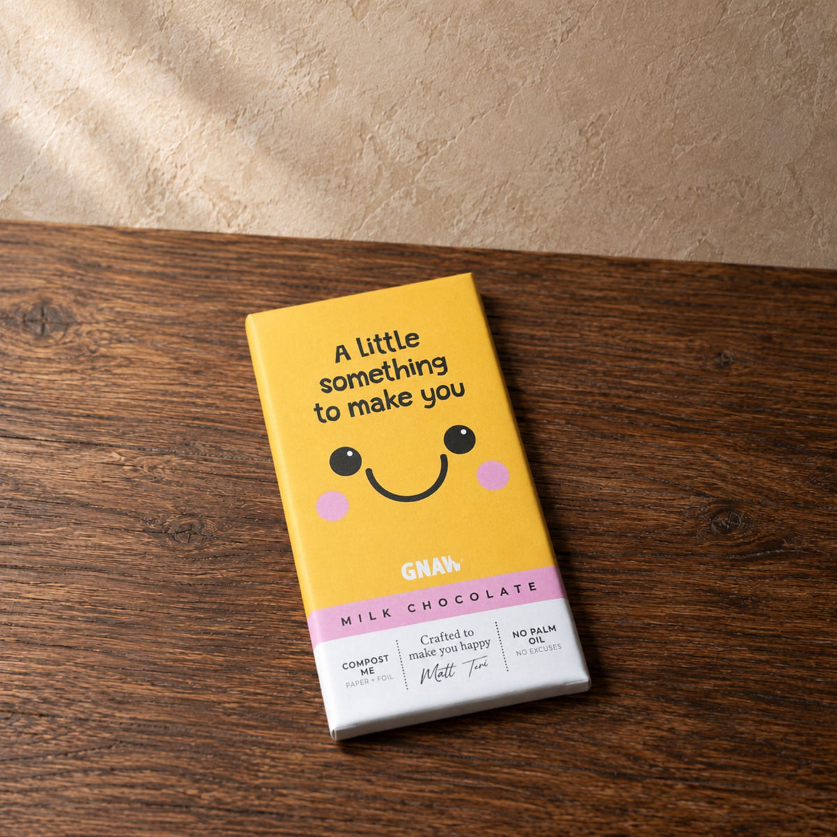 A yellow GNAW milk chocolate bar with a cute smiling face and pink cheeks, resting on a dark wooden surface with a textured beige wall in the background.
