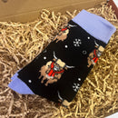 Black Christmas socks with lilac cuffs featuring Highland cow and snowflake pattern, displayed on shredded paper packaging inside a gift box.