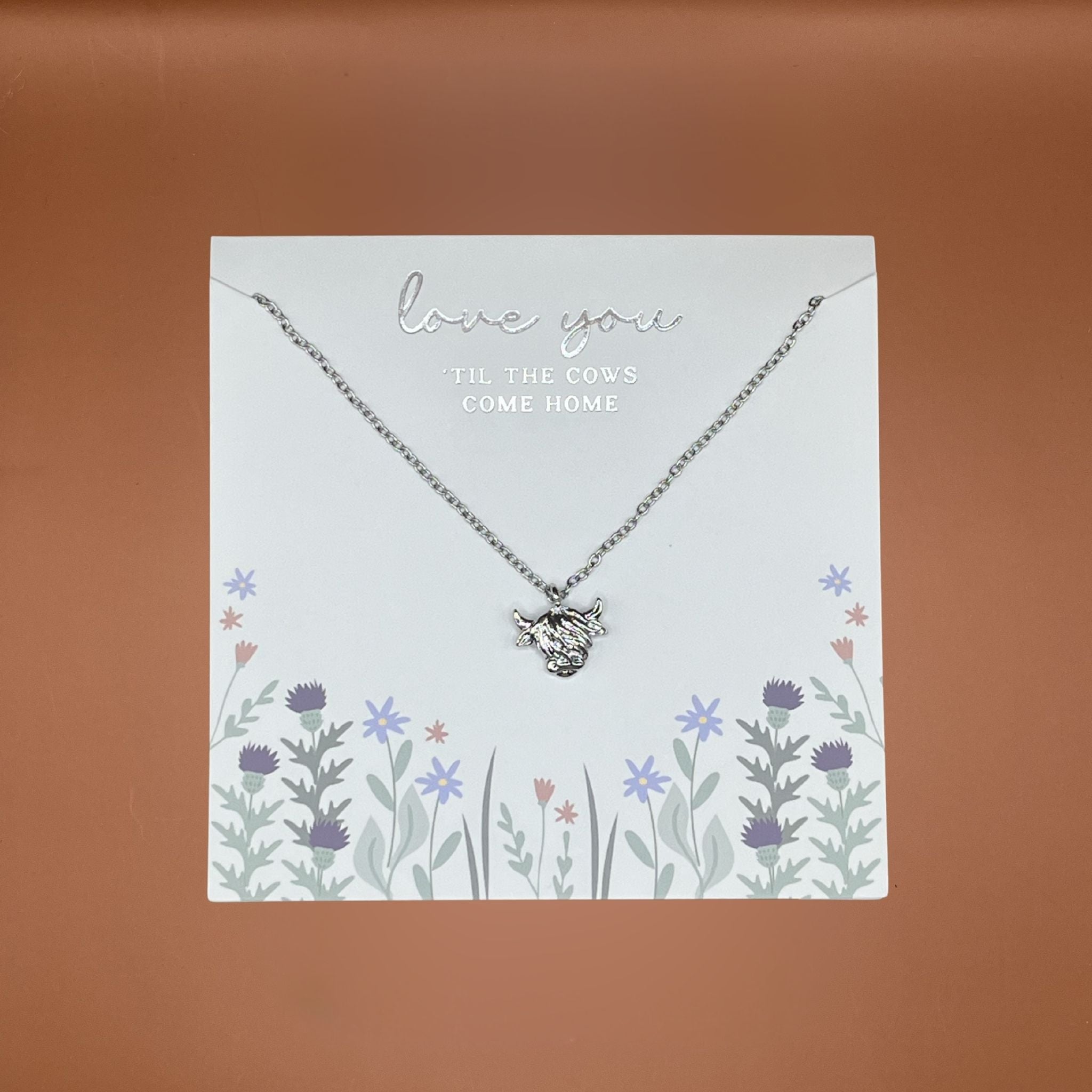 Silver necklace with a cow pendant on a decorative card with floral designs and text.