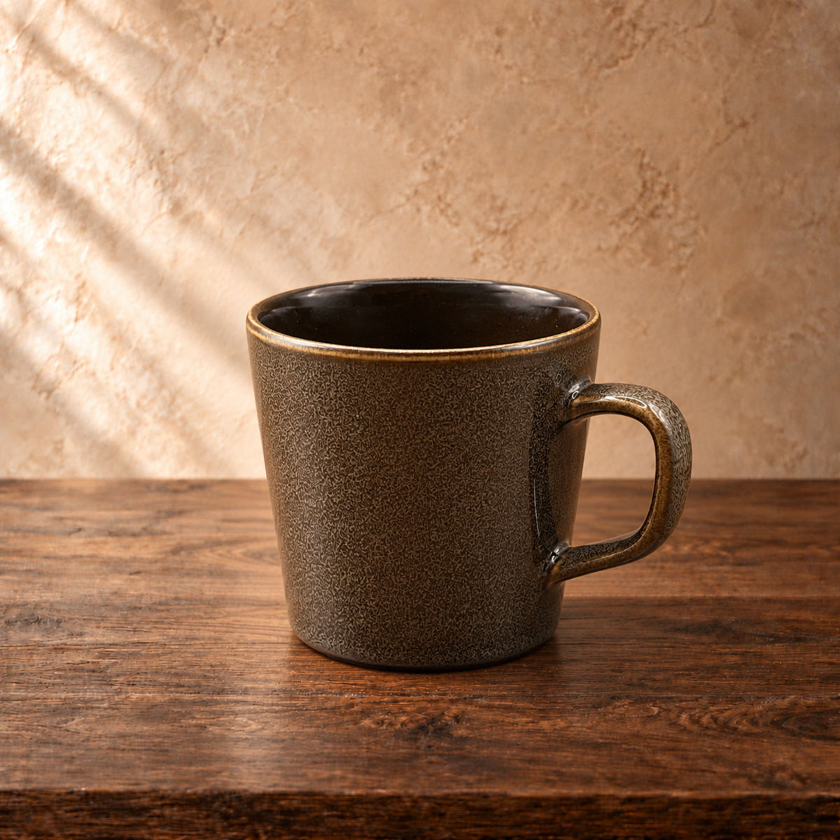 A rustic ceramic coffee mug with a speckled dark brown and black exterior and a warm gold-toned rim, sitting on a dark wooden table against a textured beige wall with soft diagonal light shadows.