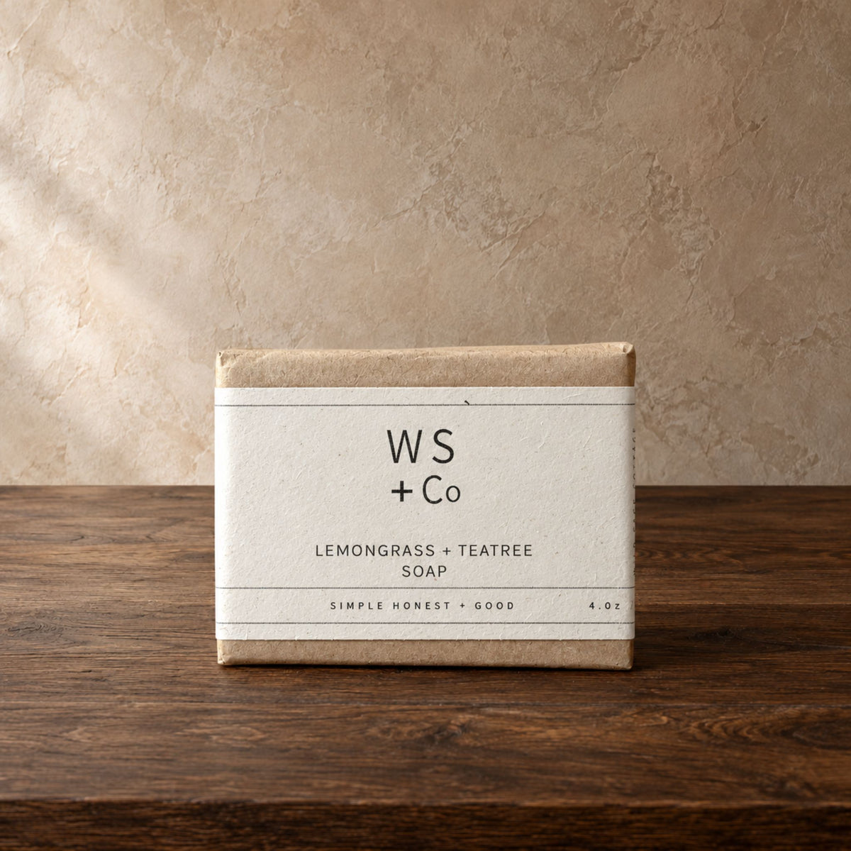 A rectangular bar of soap in a kraft paper box with a white paper label reading 'WS + Co' and 'LEMONGRASS + TEATREE SOAP', placed on a dark wooden surface against a textured beige wall.