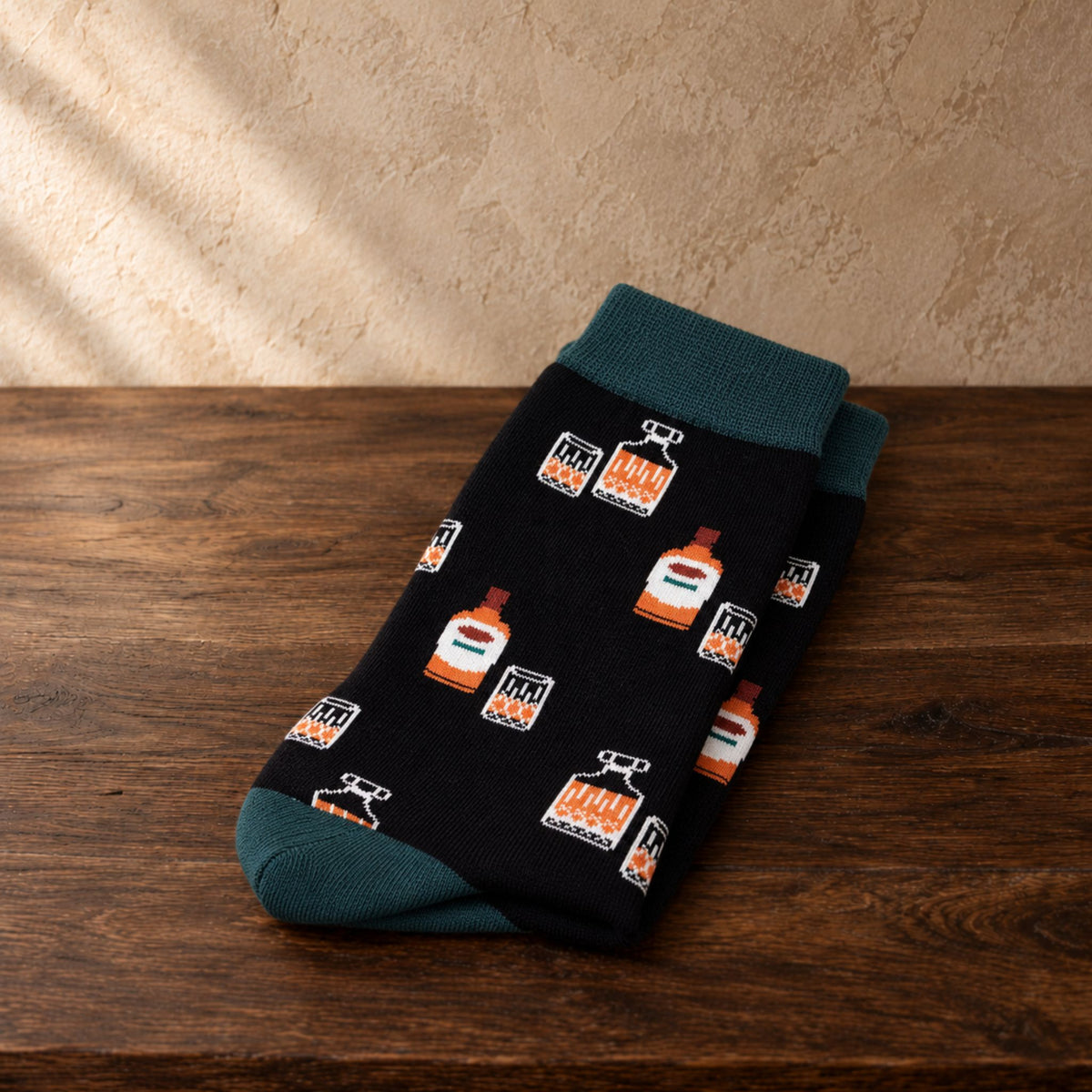 A pair of black crew socks with a repeating pattern of orange and white whiskey bottles and a dark teal cuff and toe, displayed on a dark wooden surface against a textured beige wall.