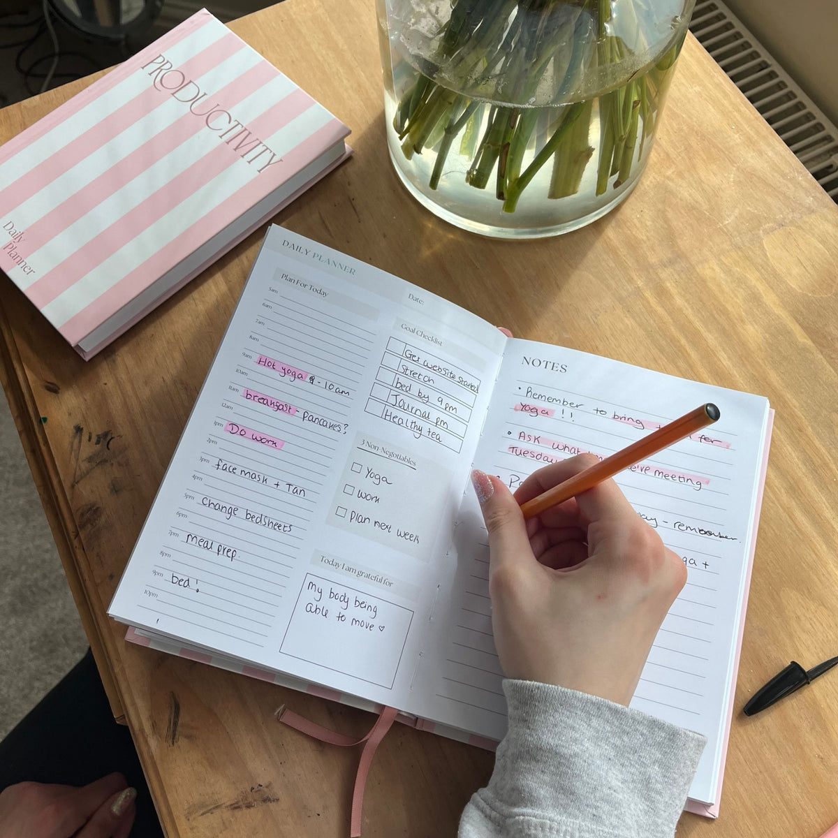 An open planner or journal on a wooden desk with a hand holding a pencil, next to a pink striped notebook and a glass vase with flowers.