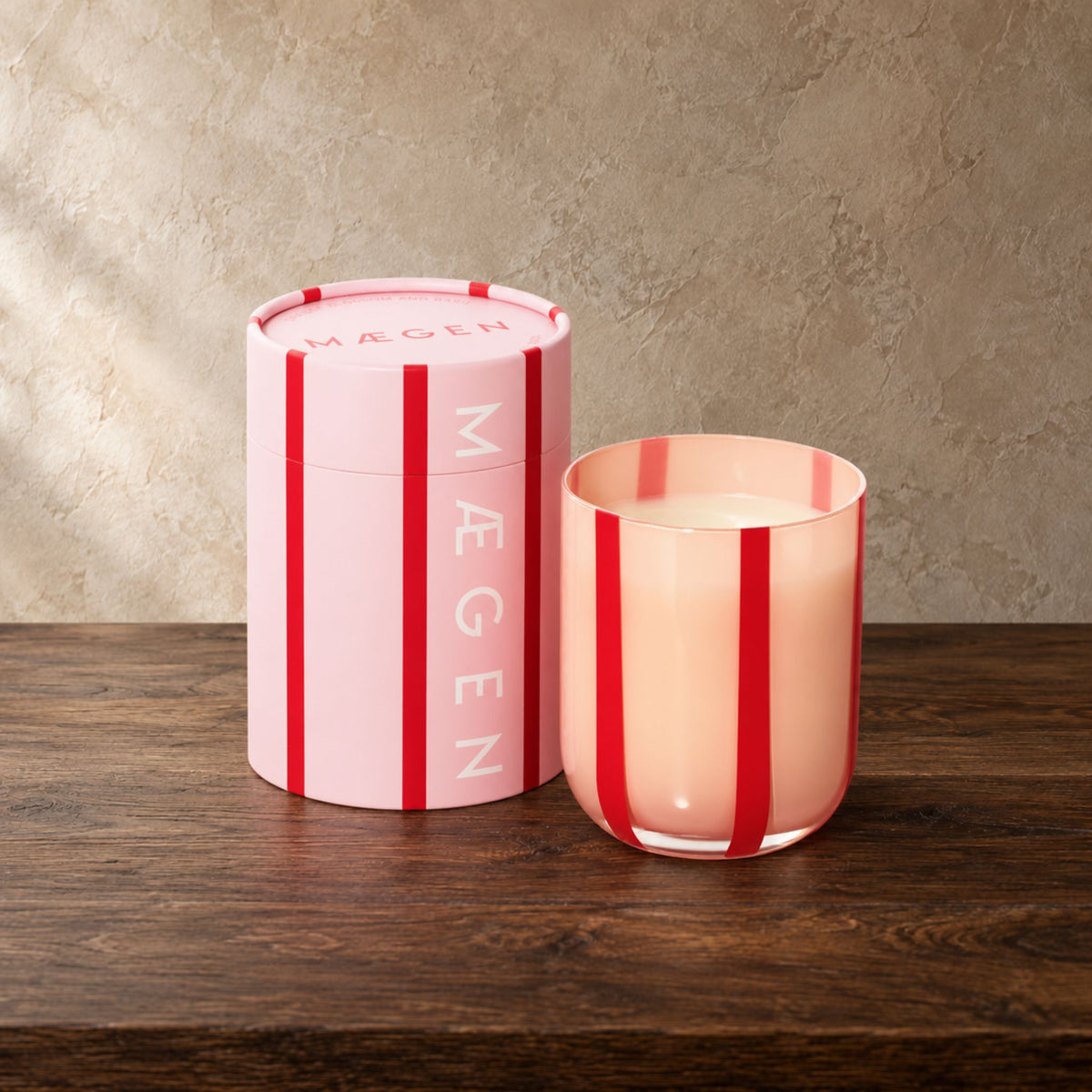 A pink and red striped candle shown with its matching pink cylindrical box on a dark wooden surface against a textured beige wall.