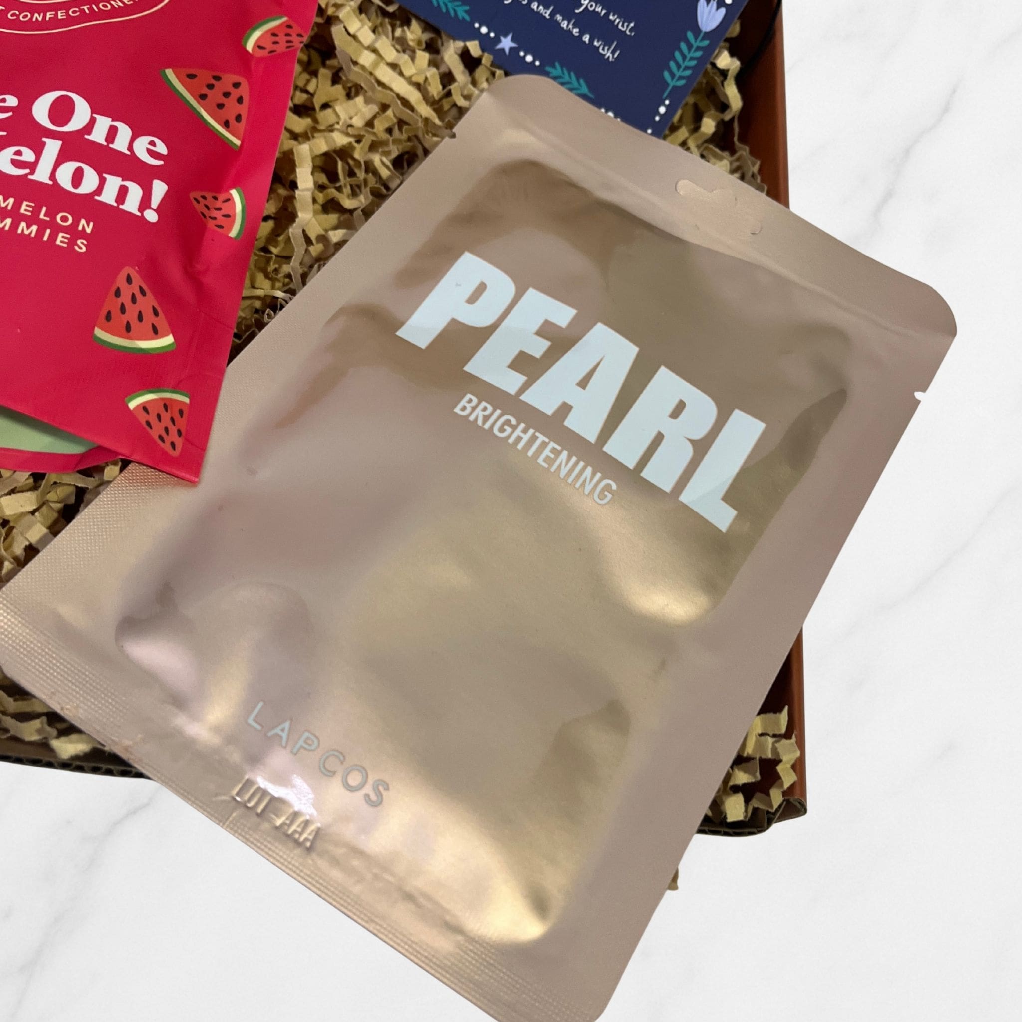 LAPCOS Pearl Brightening sheet mask in metallic packaging placed inside a shredded paper gift box beside watermelon gummies