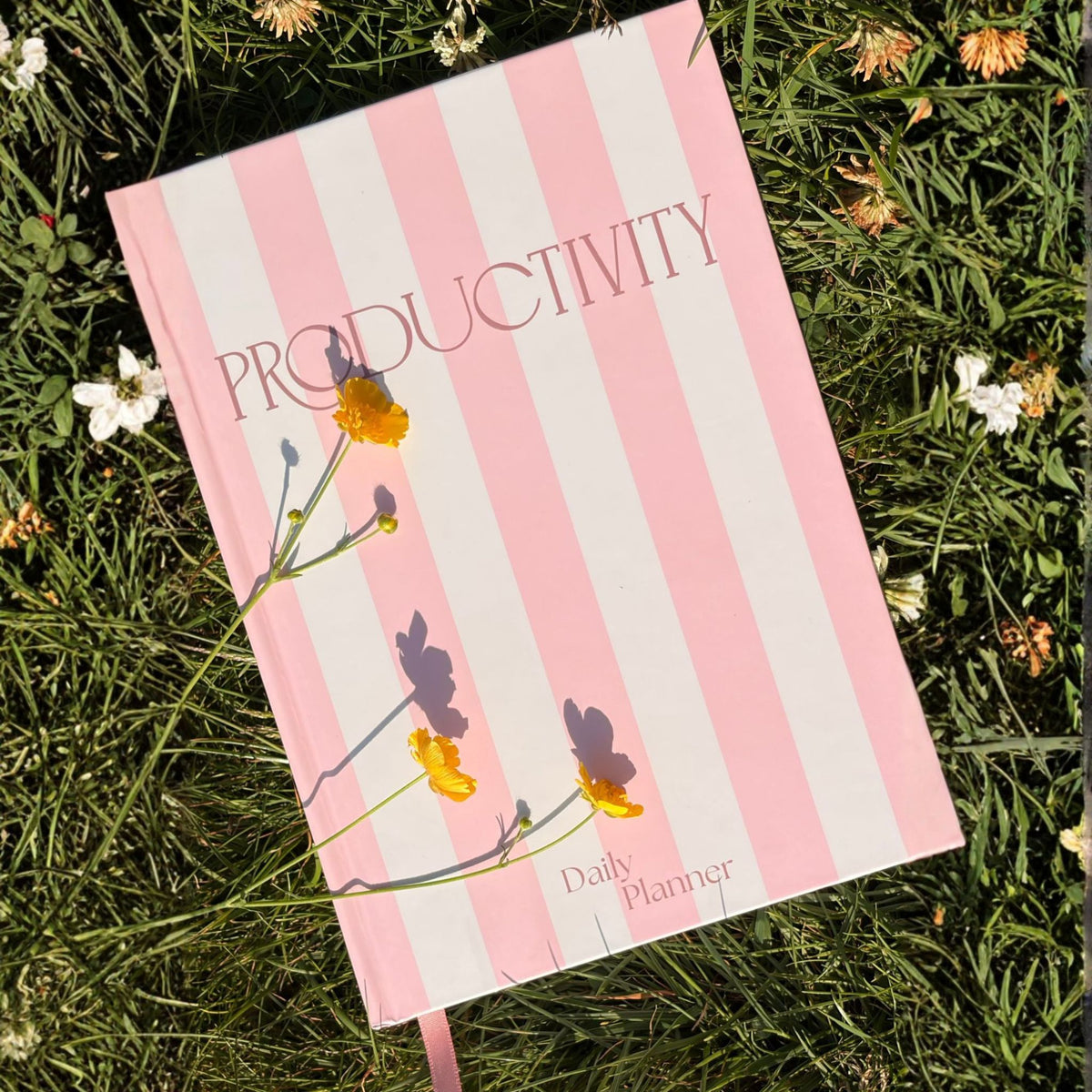 Pink and white striped productivity plannerlay on a grass field with yellow and white flowers scattered around.