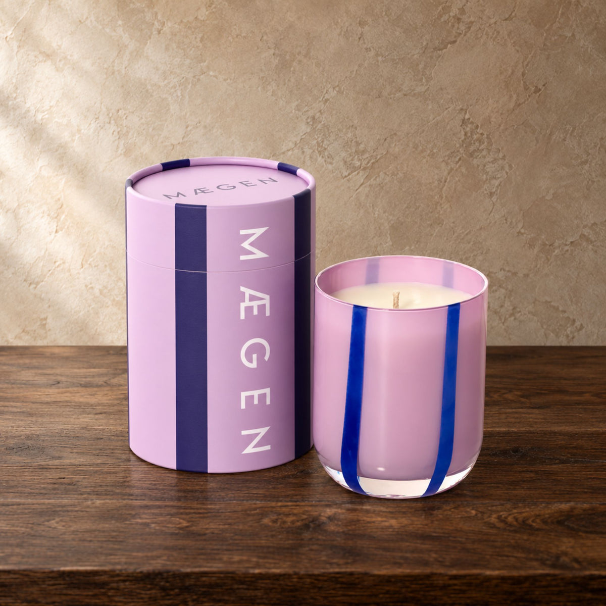 Lilac candle with dark purple strips next to matching cylinder packaging on a dark oak table in front of a beige textured wall.