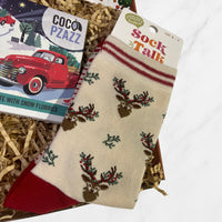 Cream Christmas socks with red toes and cuffs featuring reindeer and holly design, displayed beside festive chocolate in shredded paper packaging.