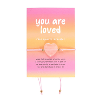 A rose quartz heart bracelet displayed on a pink and orange gradient card reading “you are loved.” The delicate pink string features gold beads, designed as a thoughtful reminder of love and positivity.