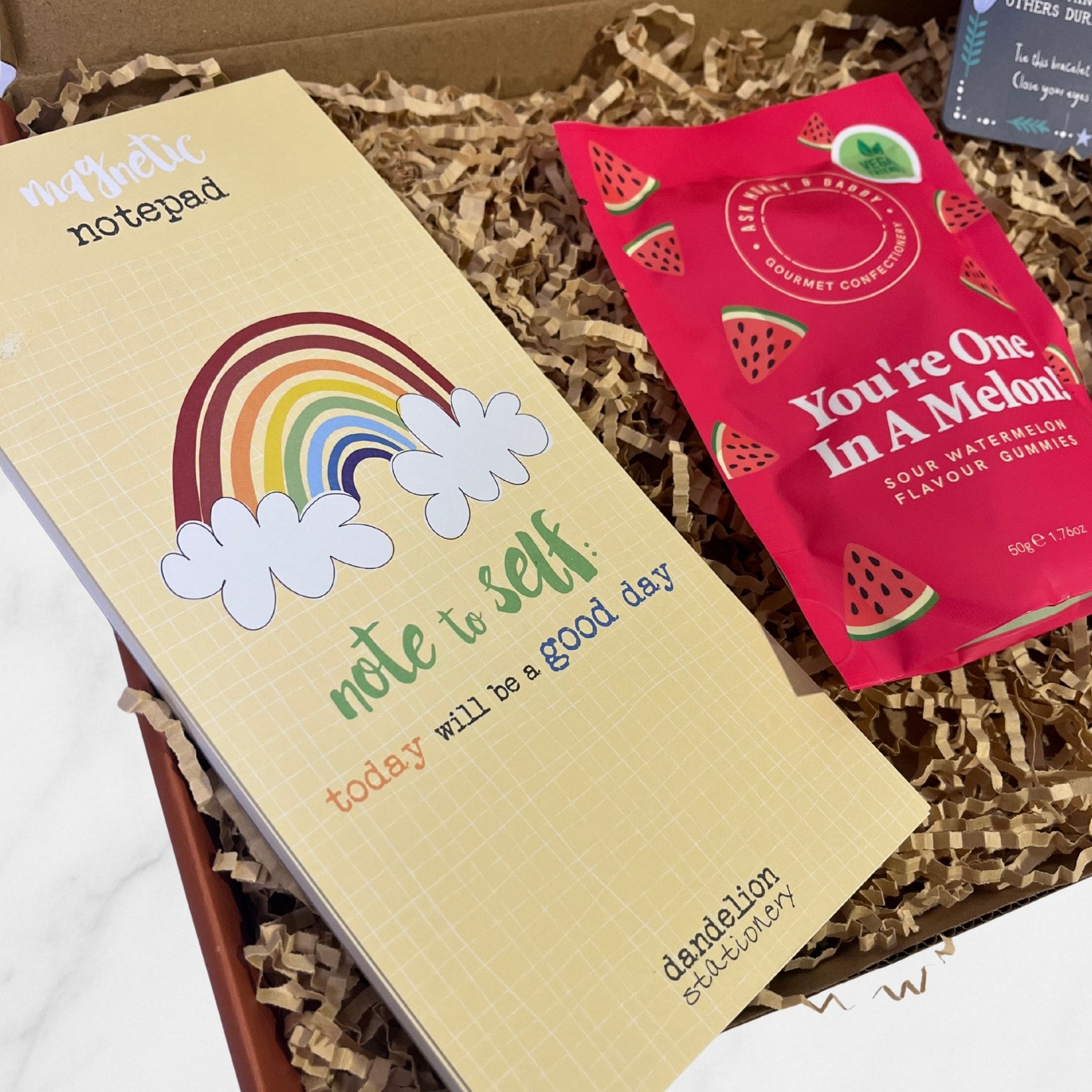Positivity-themed “Note to Self” magnetic notepad and “You’re One In A Melon” watermelon gummies displayed inside a shredded paper gift box