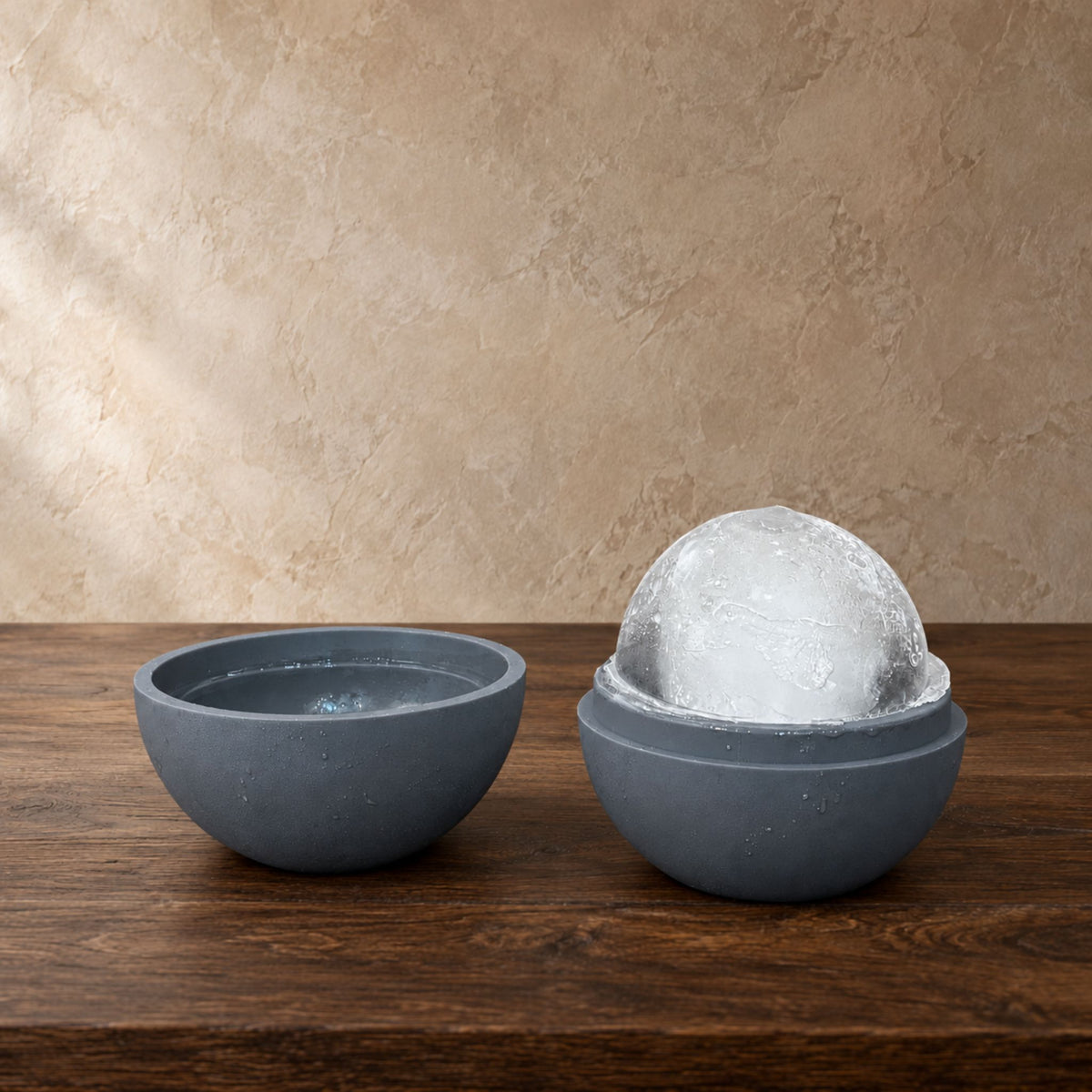 A grey silicone ice ball mold shown open on a wooden table, with one half containing an ice sphere, against a textured beige wall.