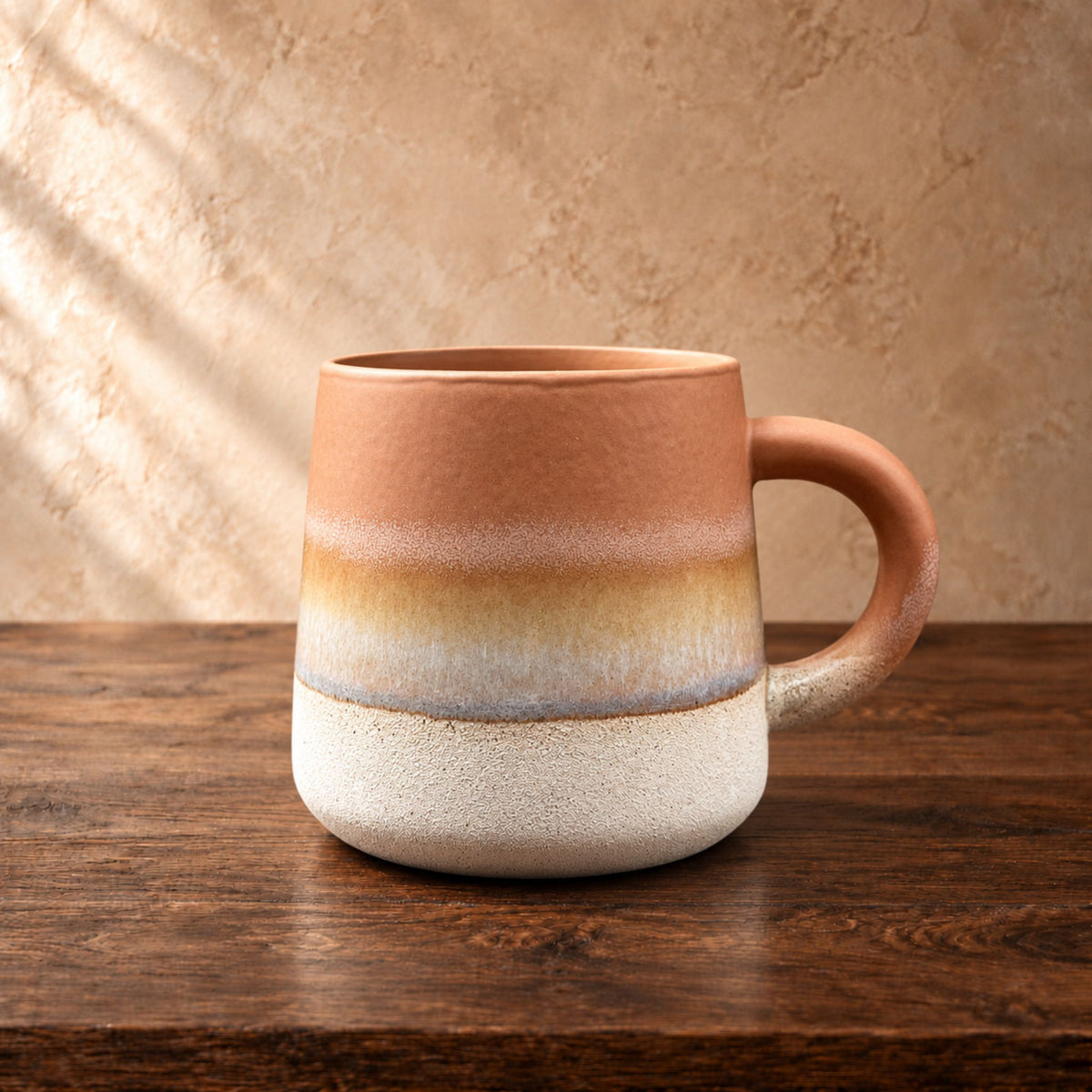 A handmade ceramic mug with a terracotta-colored top half, a middle band of beige and light blue-gray, and a textured off-white bottom half, sitting on a dark wooden table against a textured beige.