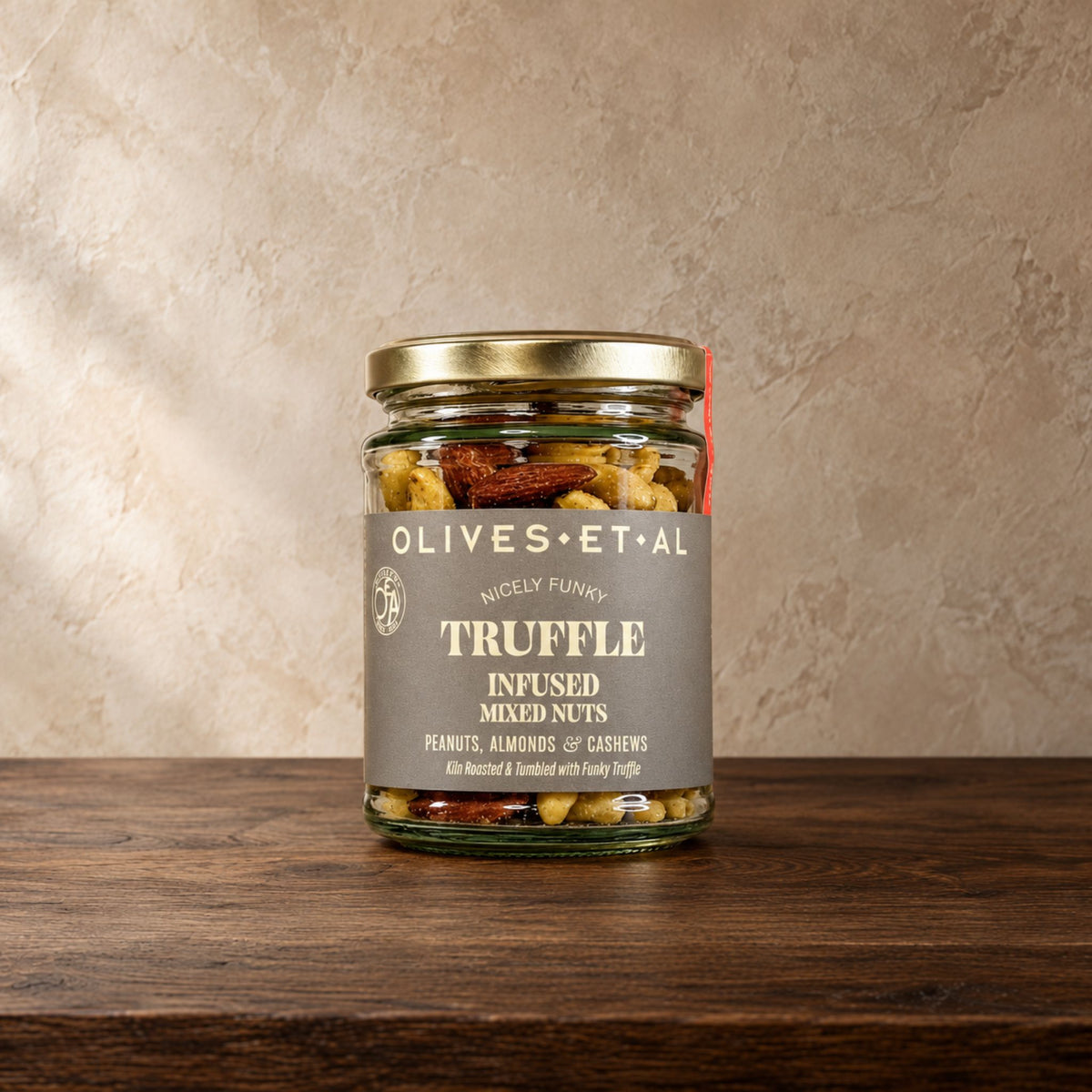 A glass jar of Olives Et Al Truffle Infused Mixed Nuts sits on a dark wooden table against a textured beige wall. The jar has a silver lid and a grey label with white text.