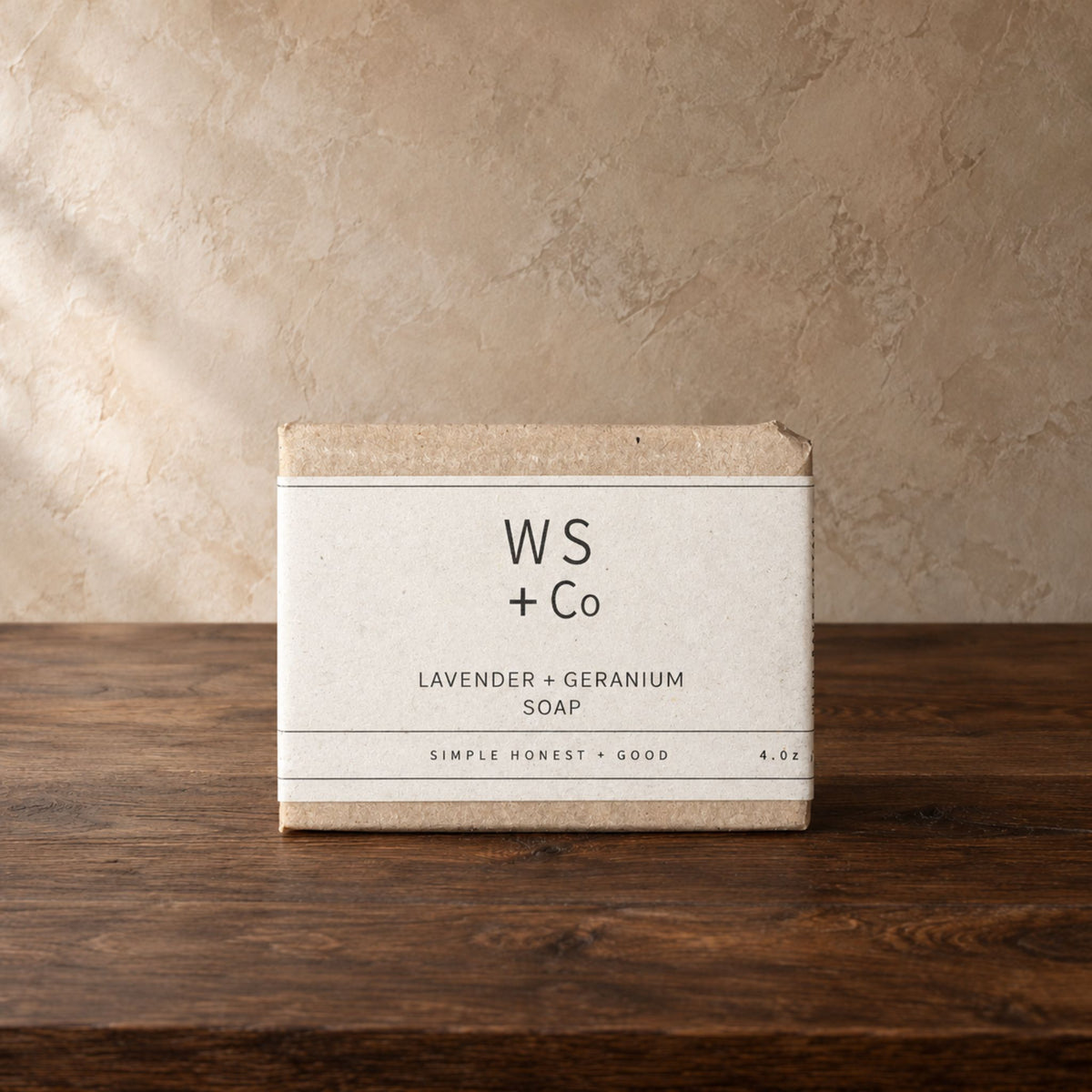 A rectangular bar of soap in a kraft paper box with a cream paper label reading 'WS + Co' and 'LAVENDER + GERANIUM SOAP', placed on a dark wooden surface against a textured beige wall.